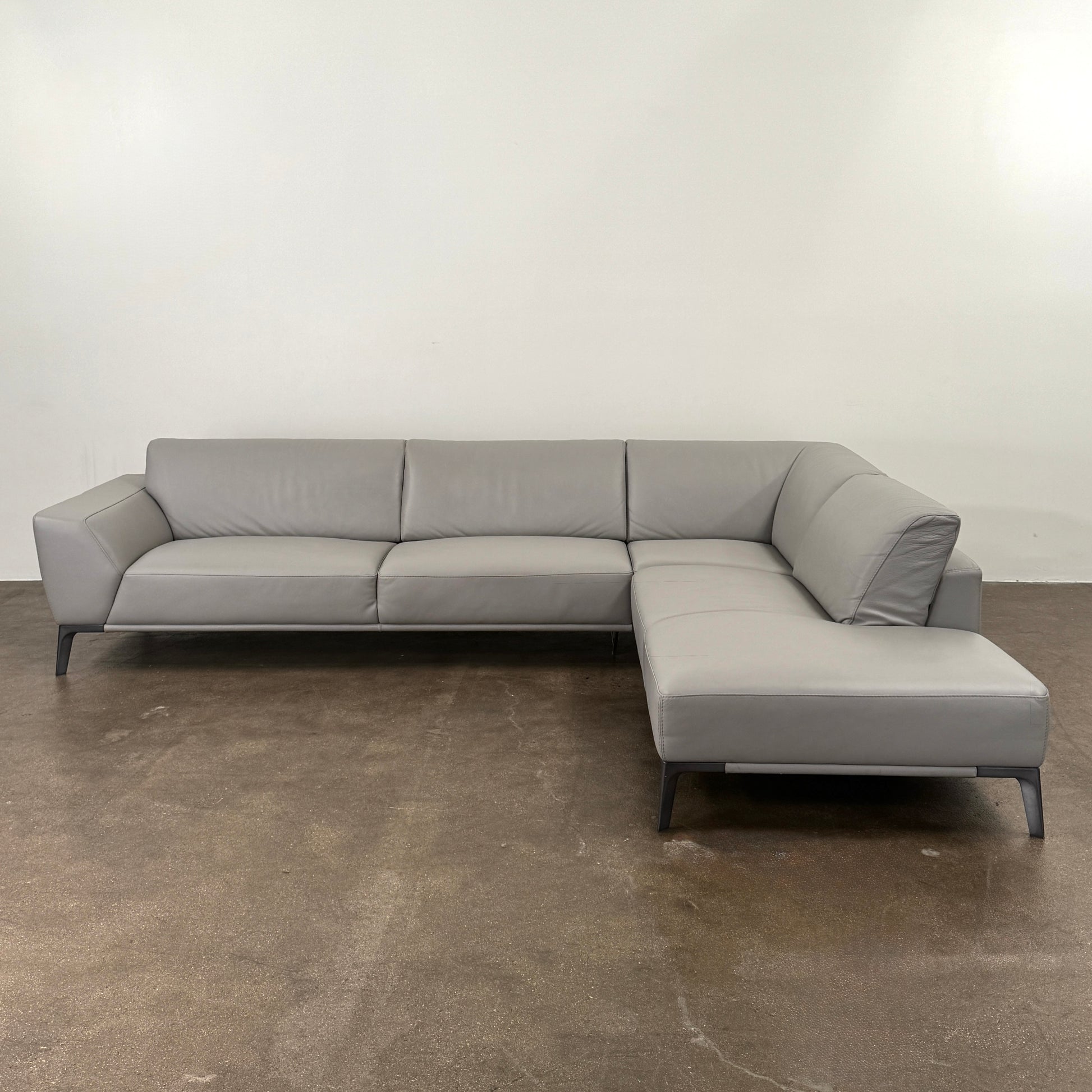 A Roche Bobois Accord Gray Leather Sectional sits in a spacious room with floor-to-ceiling windows and light wood floors, overlooking a cityscape.
