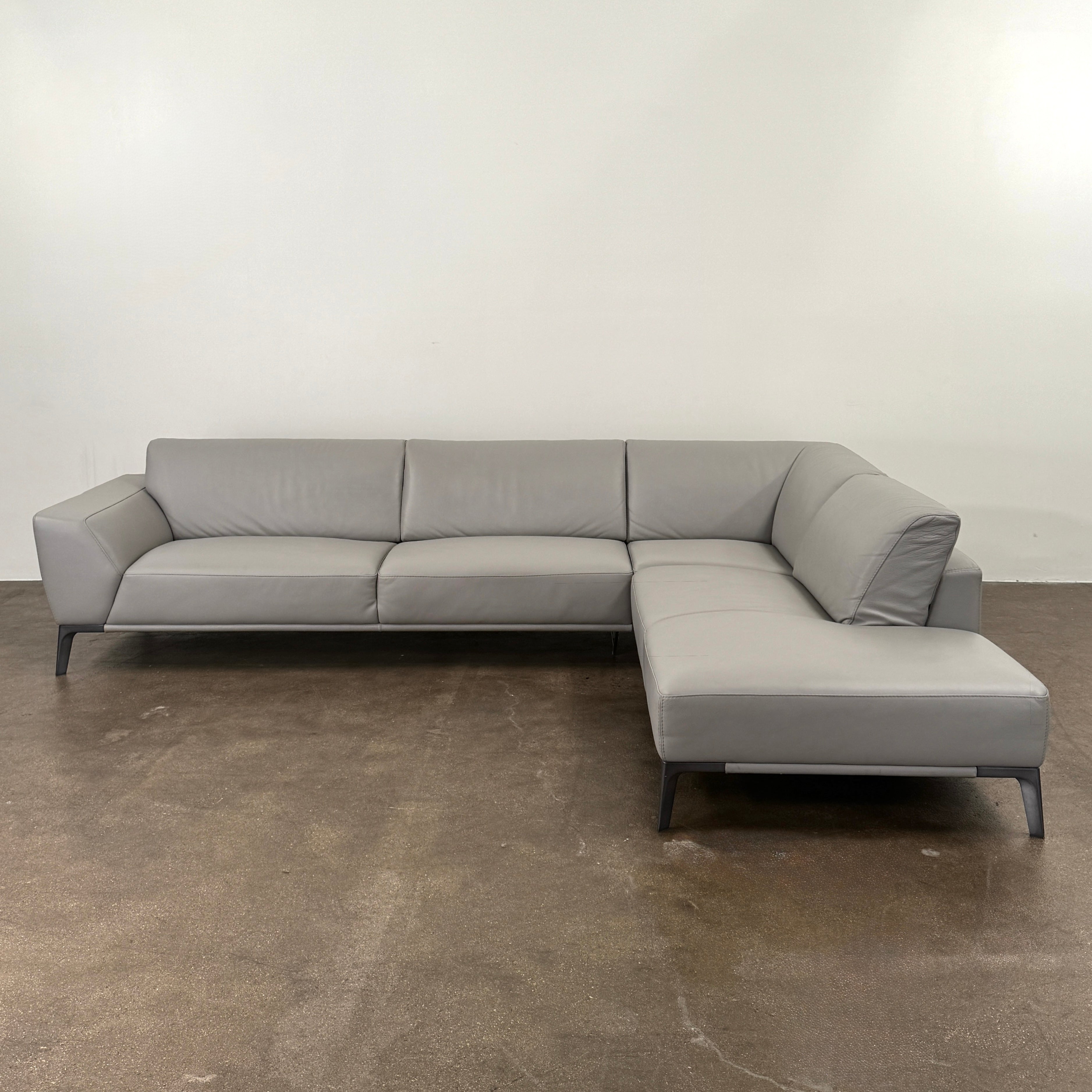 A Roche Bobois Accord Gray Leather Sectional sits in a spacious room with floor-to-ceiling windows and light wood floors, overlooking a cityscape.