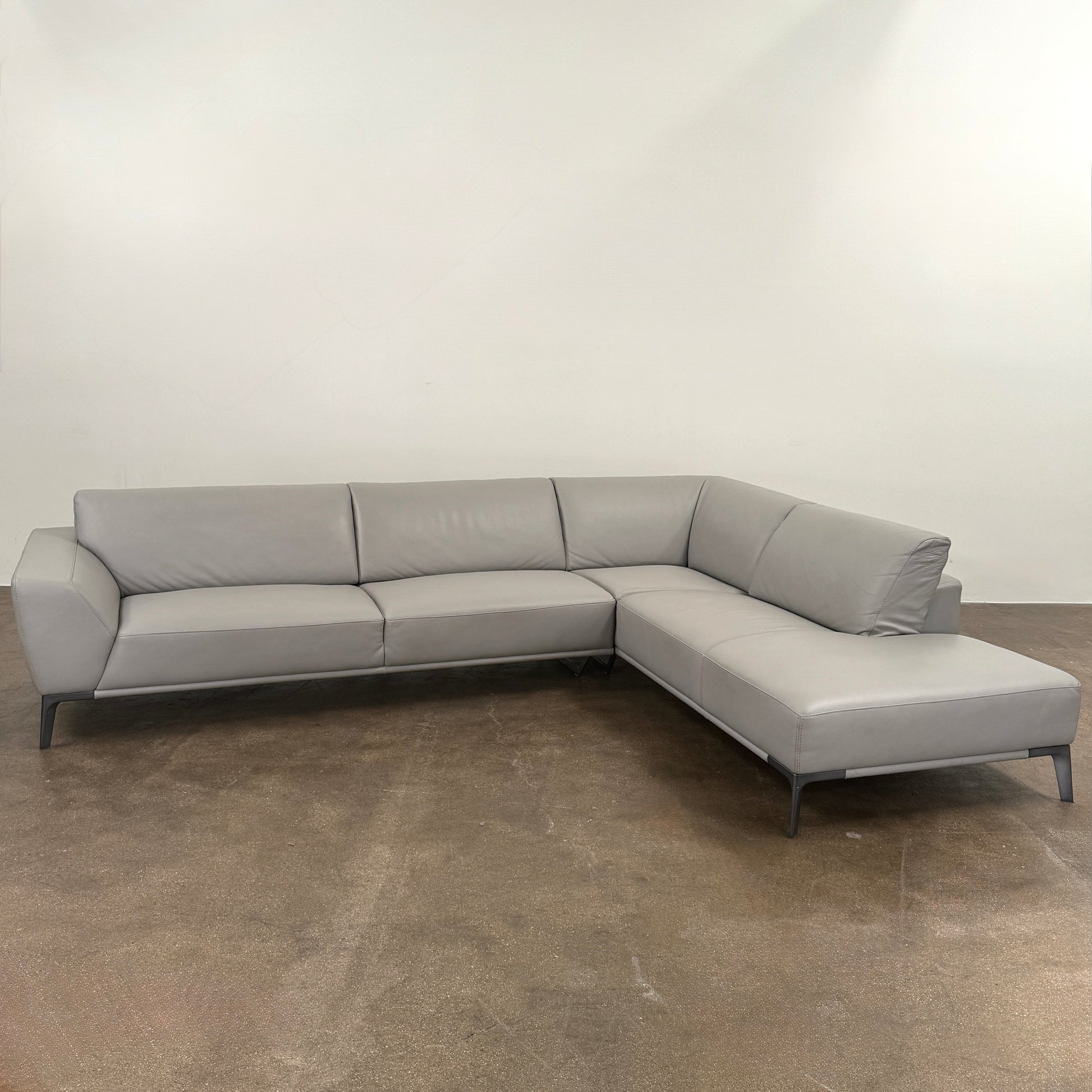 A Roche Bobois Accord Gray Leather Sectional sits in a spacious room with floor-to-ceiling windows and light wood floors, overlooking a cityscape.