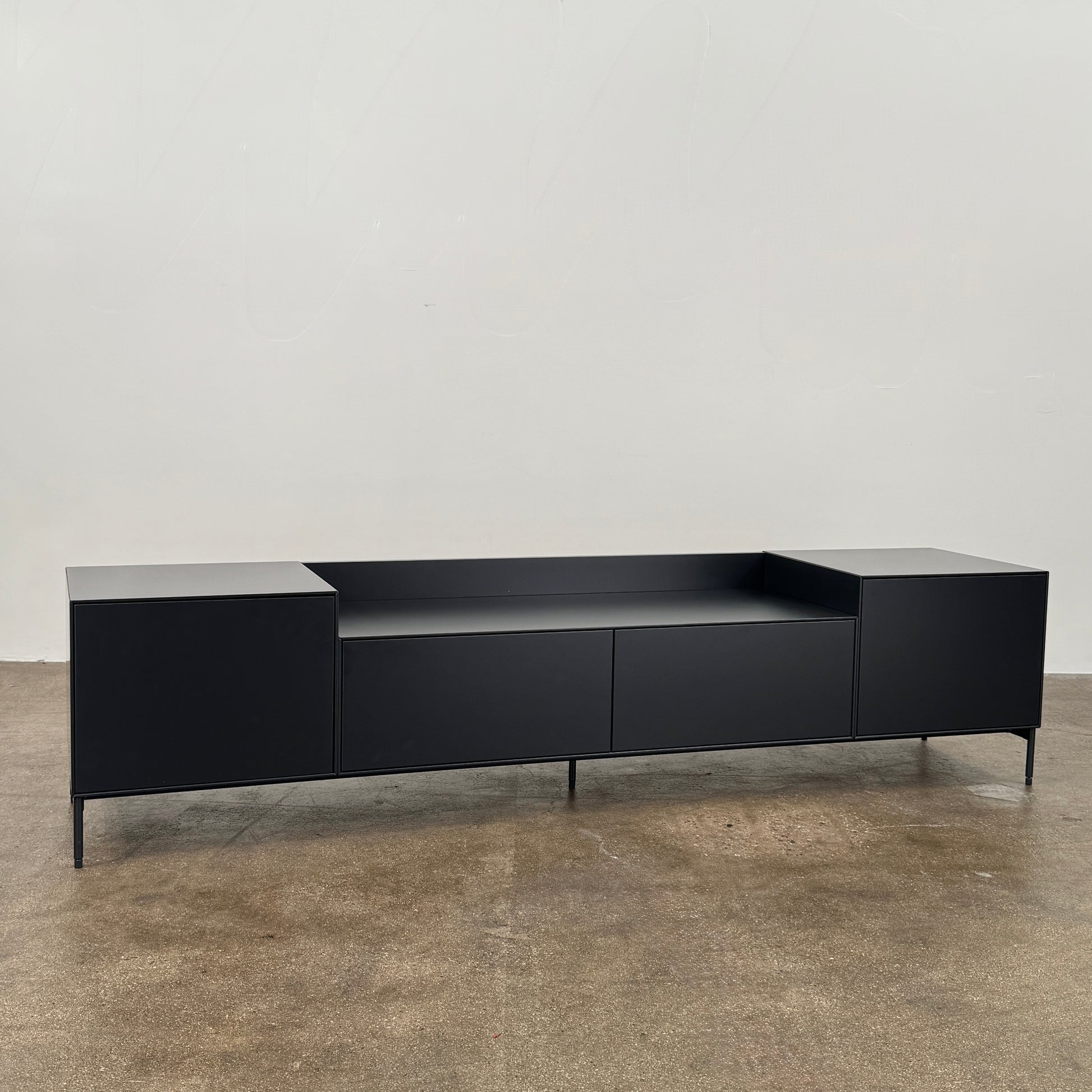 The Treku Lauki Media Unit by Treku features a black minimalist design with two closed side cabinets, two central drawers, an open shelf, and built-in cord management on slim metal legs—perfect for modern spaces.