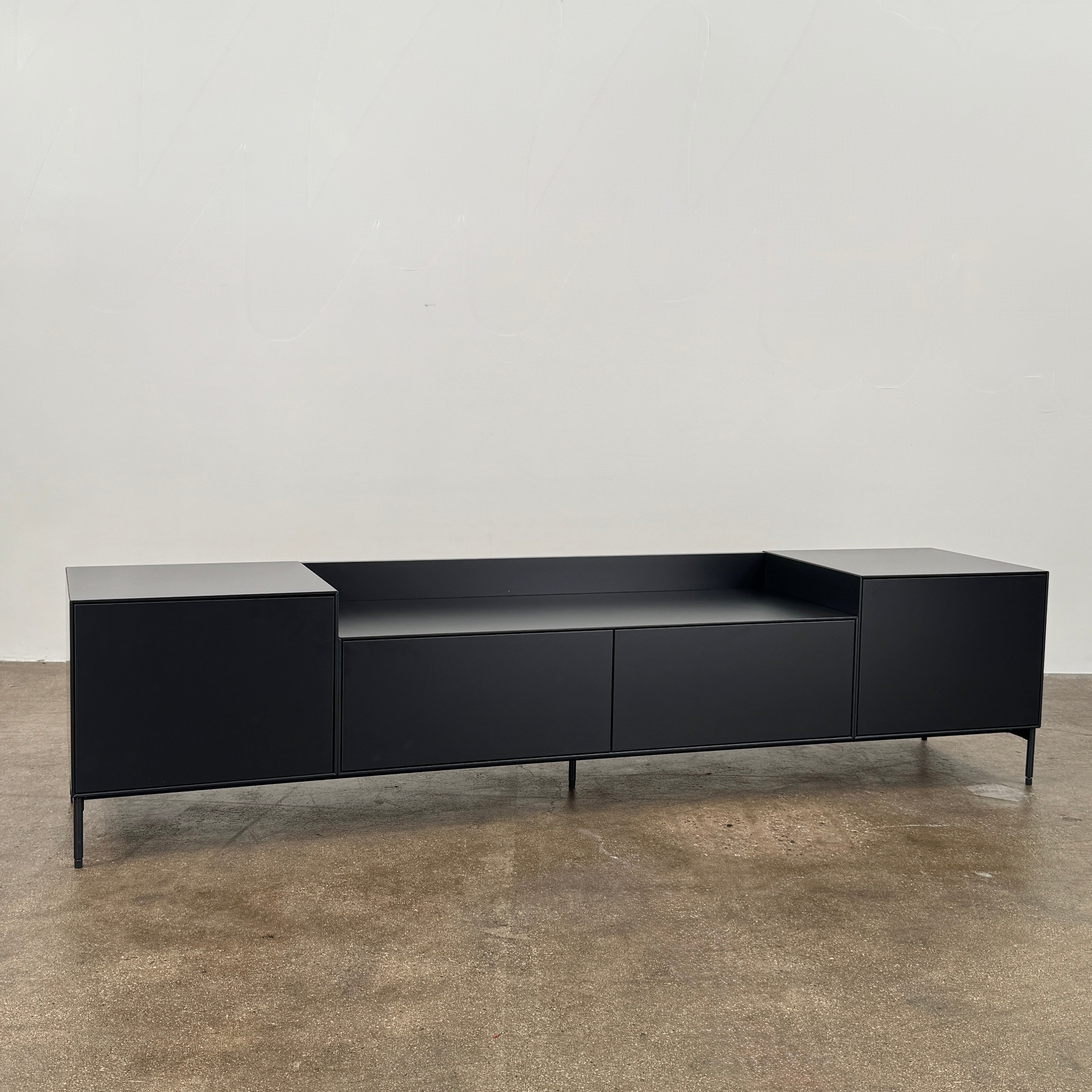 The Treku Lauki Media Unit by Treku features a black minimalist design with two closed side cabinets, two central drawers, an open shelf, and built-in cord management on slim metal legs—perfect for modern spaces.