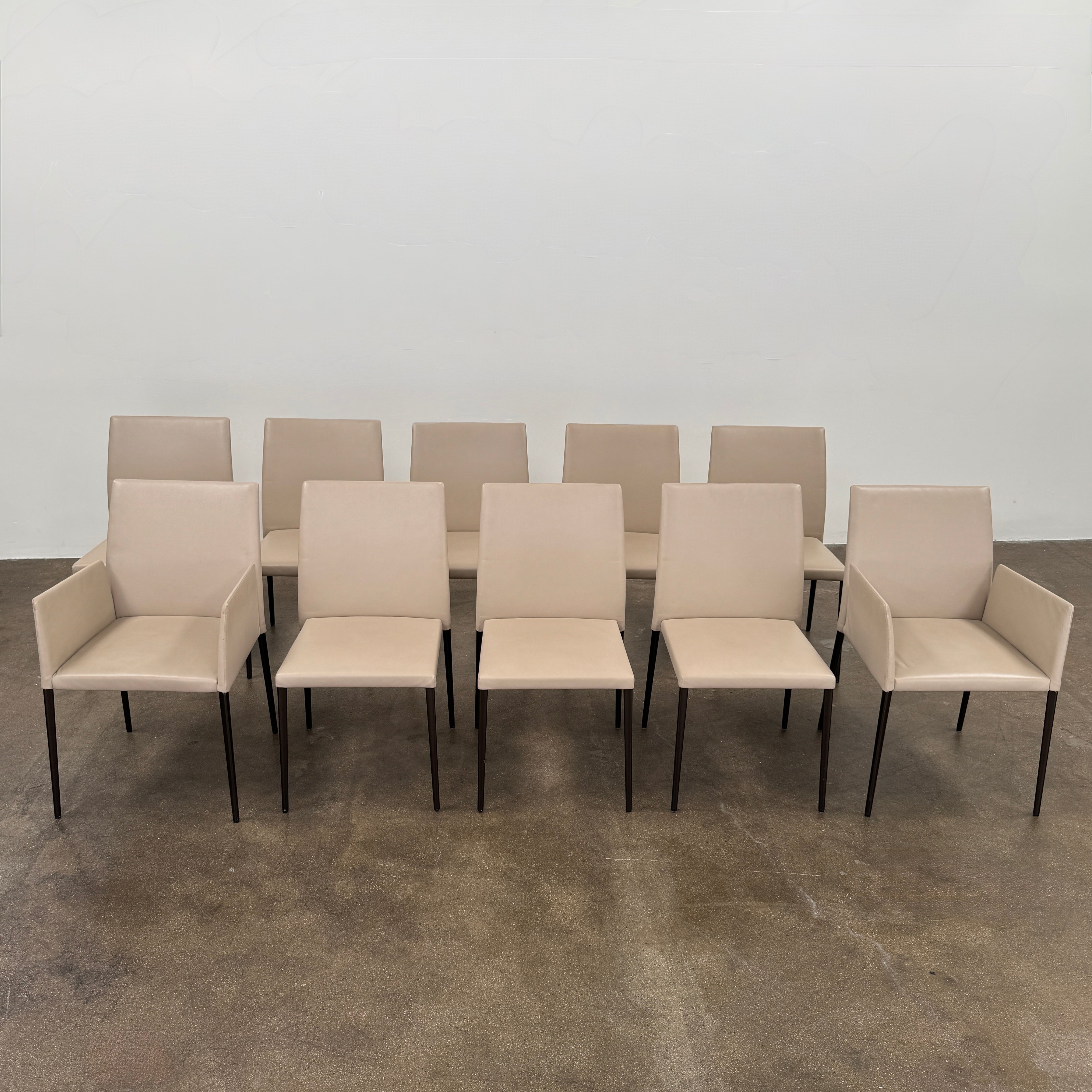 A set of Walter Knoll Deen Dining Chairs (2 armchairs, 8 side chairs) by Walter Knoll with beige upholstery and black legs are arranged in three rows on a brown concrete floor against a plain white wall.