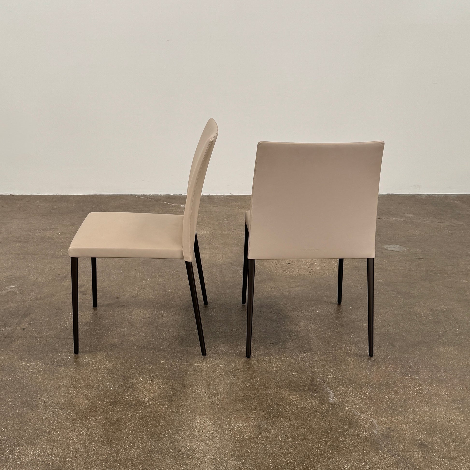 A set of Walter Knoll Deen Dining Chairs (2 armchairs, 8 side chairs) by Walter Knoll with beige upholstery and black legs are arranged in three rows on a brown concrete floor against a plain white wall.