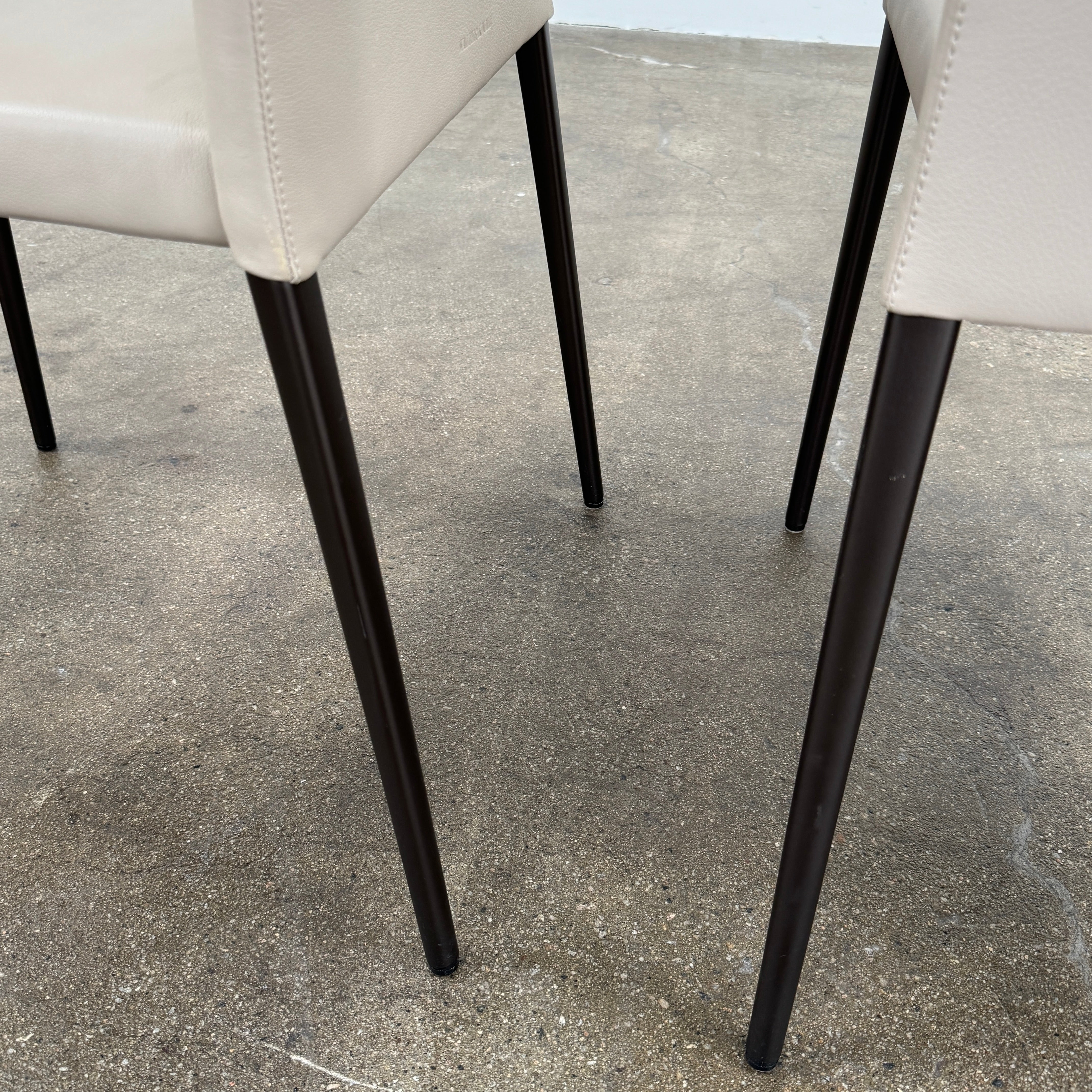 A set of Walter Knoll Deen Dining Chairs (2 armchairs, 8 side chairs) by Walter Knoll with beige upholstery and black legs are arranged in three rows on a brown concrete floor against a plain white wall.