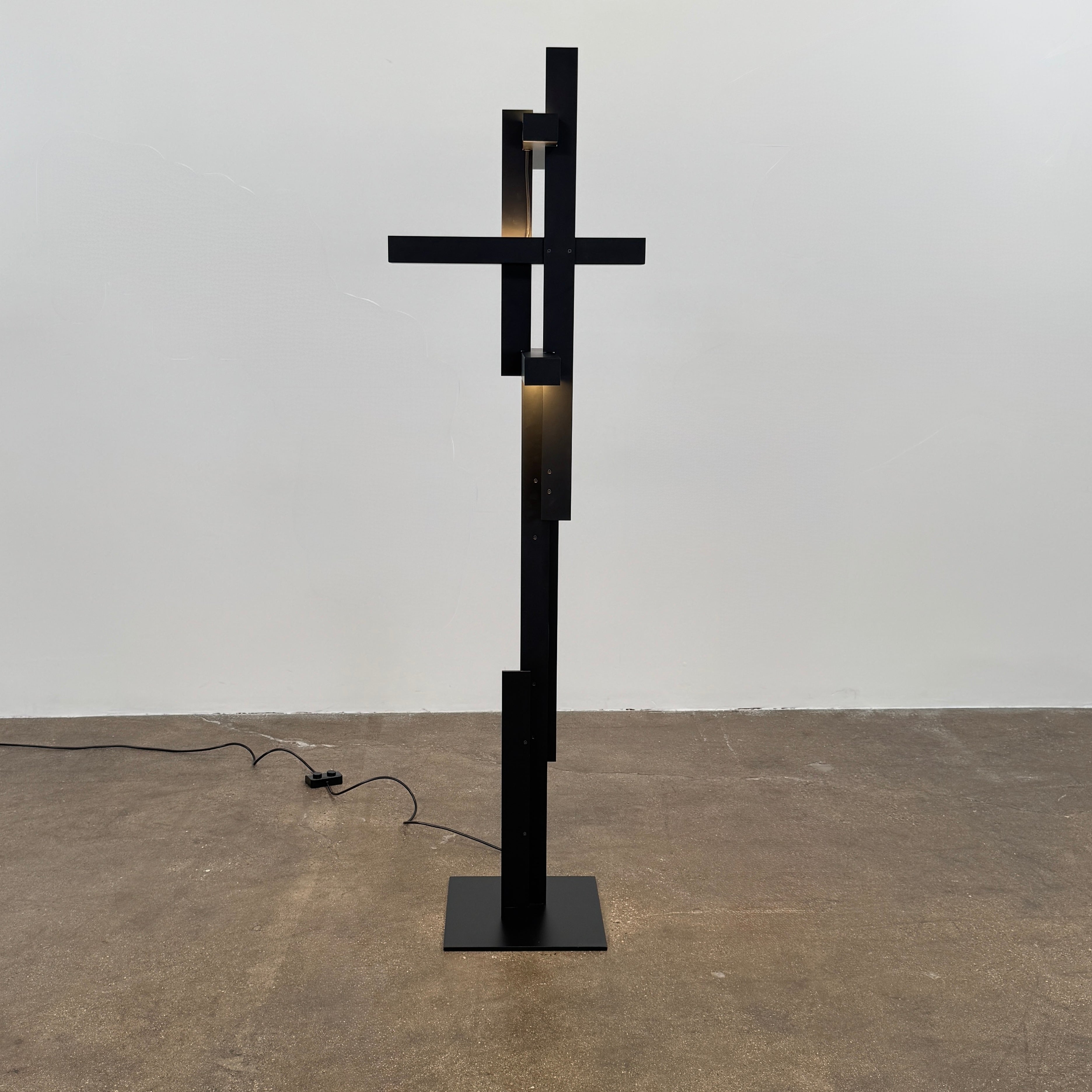 The Oluce LAS 376 LED Floor Lamp by Oluce features intersecting black rectangular panels on a flat base, with a visible power cord. Its sculptural design makes it a striking contemporary statement piece for any space.