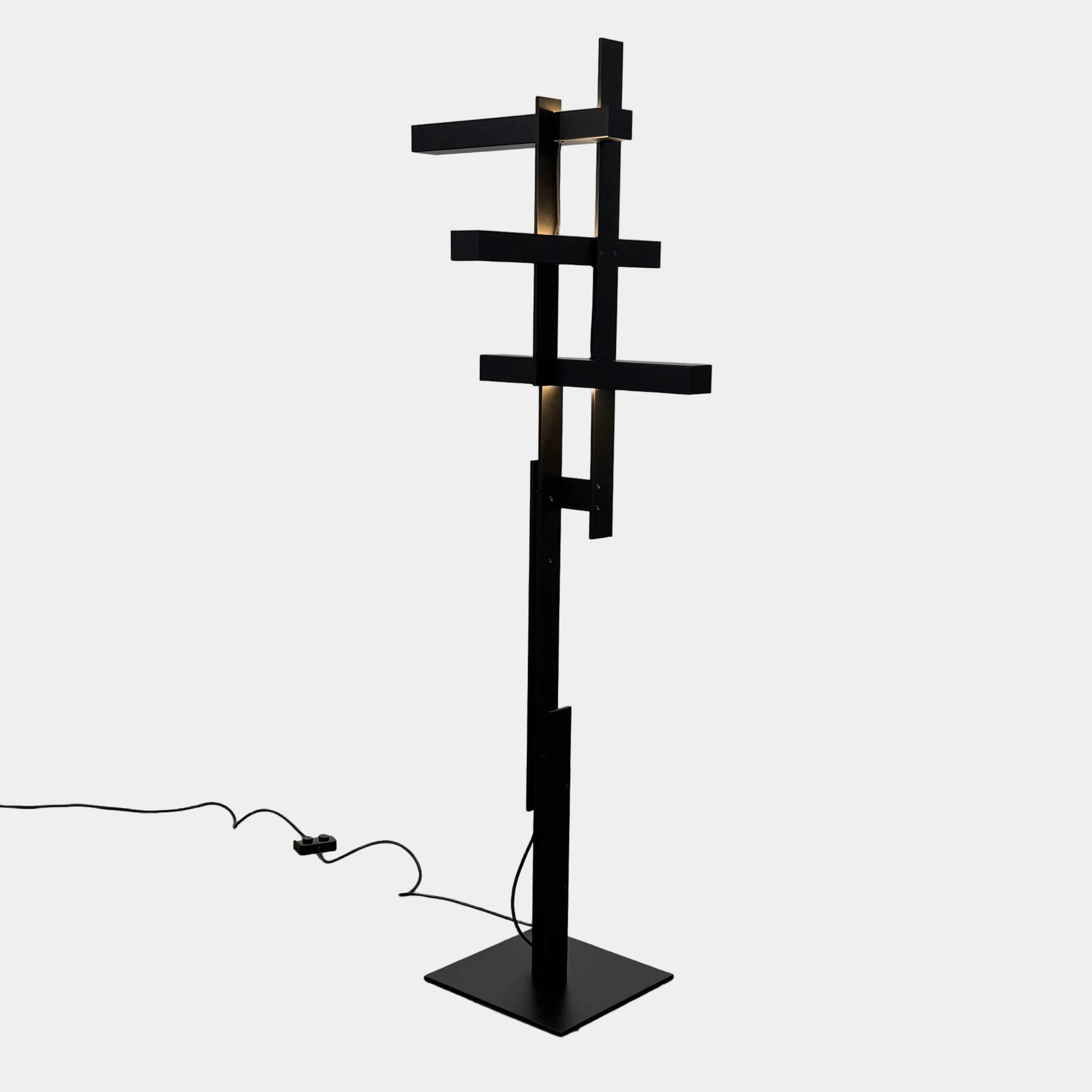 The Oluce LAS 376 LED Floor Lamp by Oluce features intersecting black rectangular panels on a flat base, with a visible power cord. Its sculptural design makes it a striking contemporary statement piece for any space.