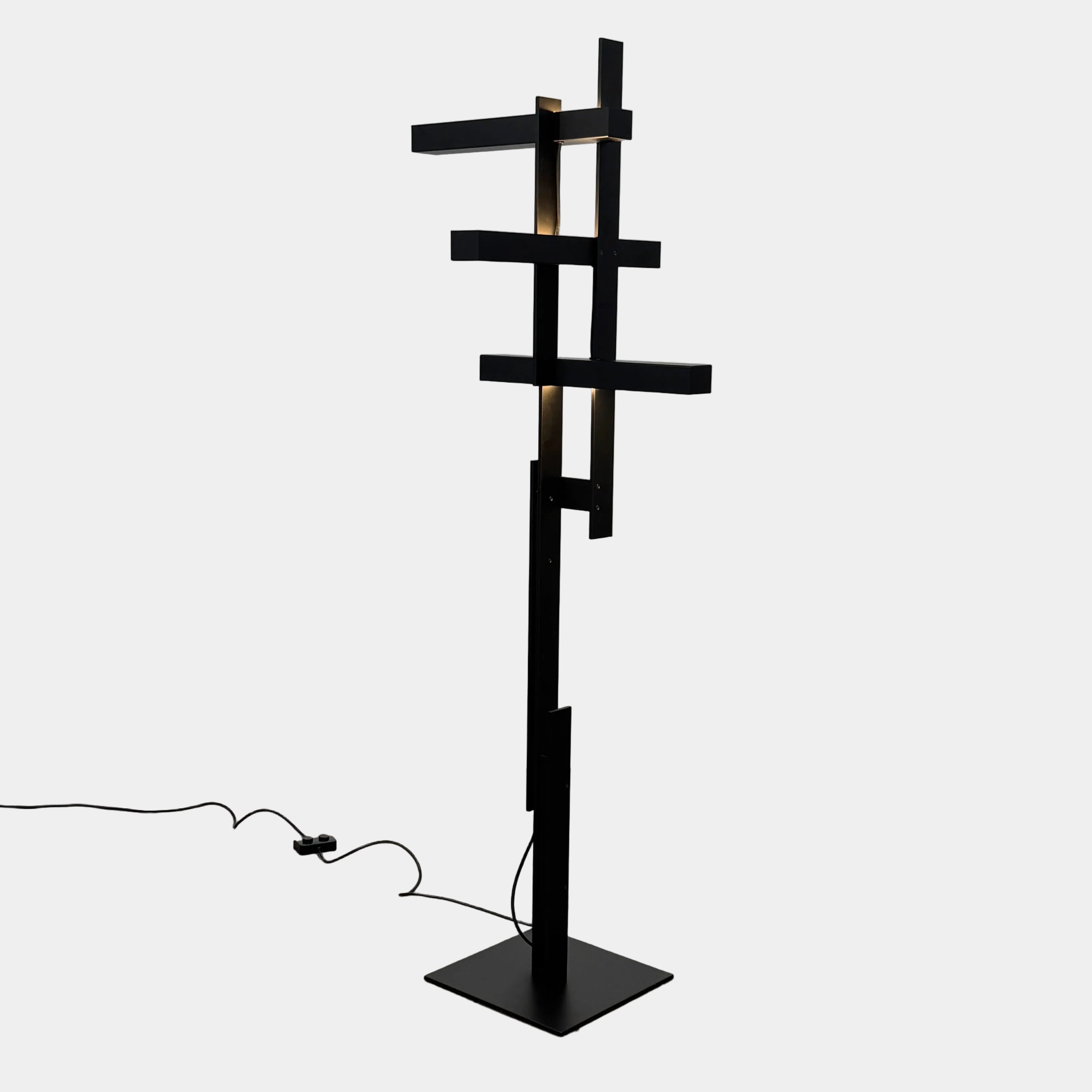 The Oluce LAS 376 LED Floor Lamp by Oluce features intersecting black rectangular panels on a flat base, with a visible power cord. Its sculptural design makes it a striking contemporary statement piece for any space.