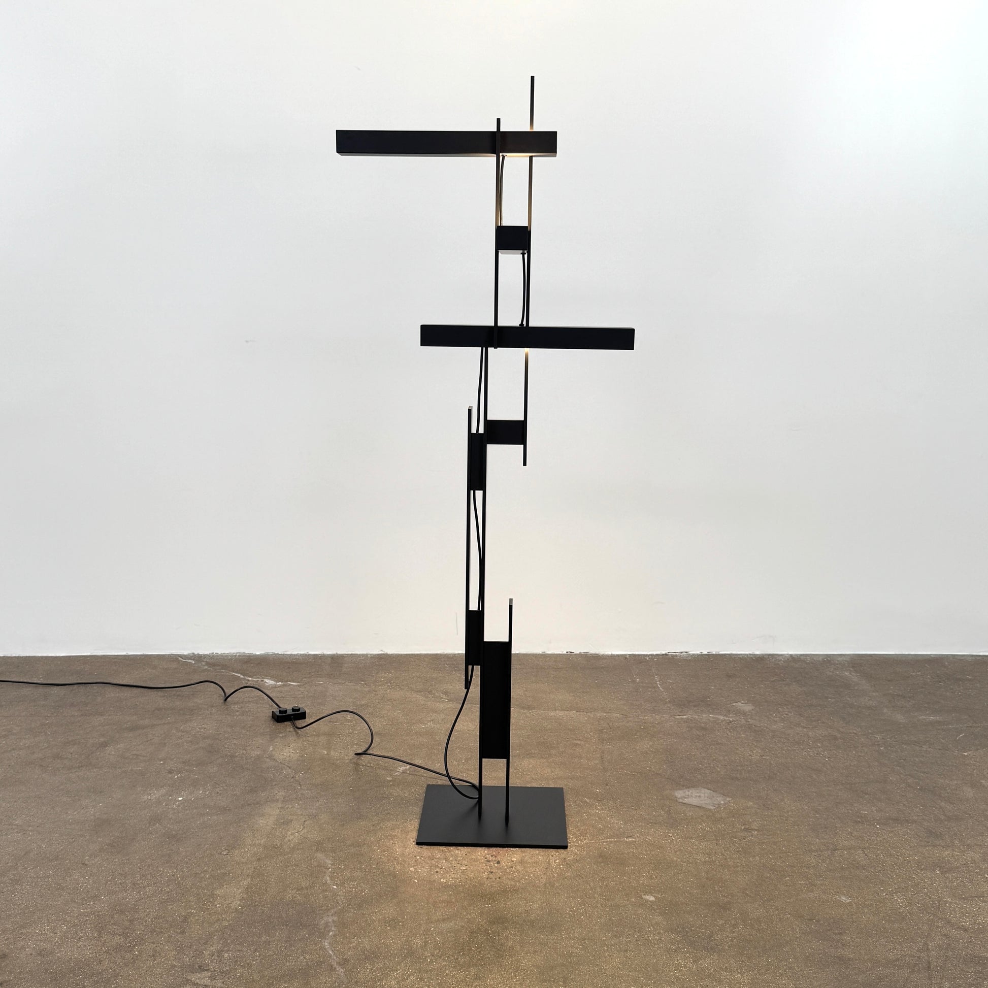 The Oluce LAS 376 LED Floor Lamp by Oluce features intersecting black rectangular panels on a flat base, with a visible power cord. Its sculptural design makes it a striking contemporary statement piece for any space.