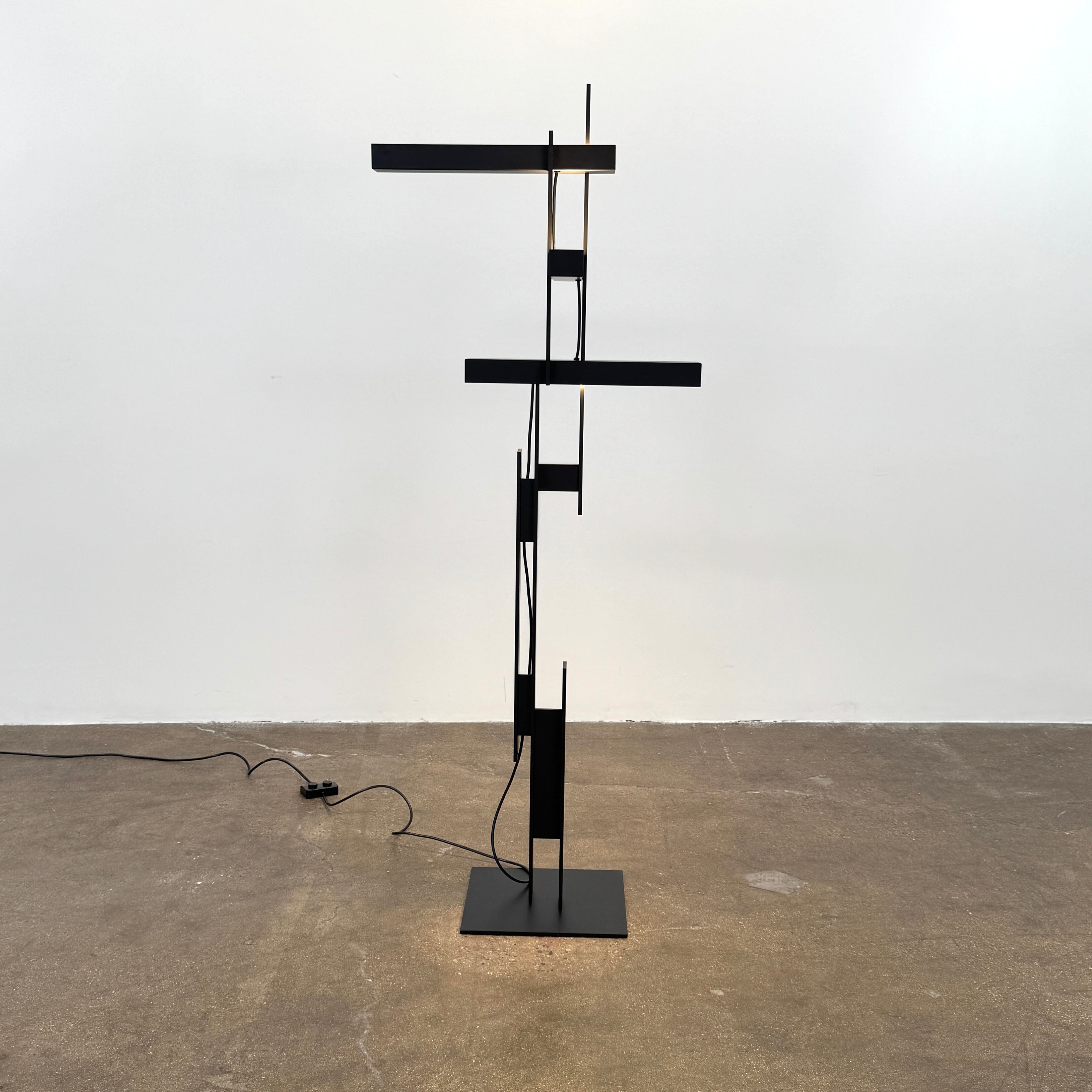 The Oluce LAS 376 LED Floor Lamp by Oluce features intersecting black rectangular panels on a flat base, with a visible power cord. Its sculptural design makes it a striking contemporary statement piece for any space.