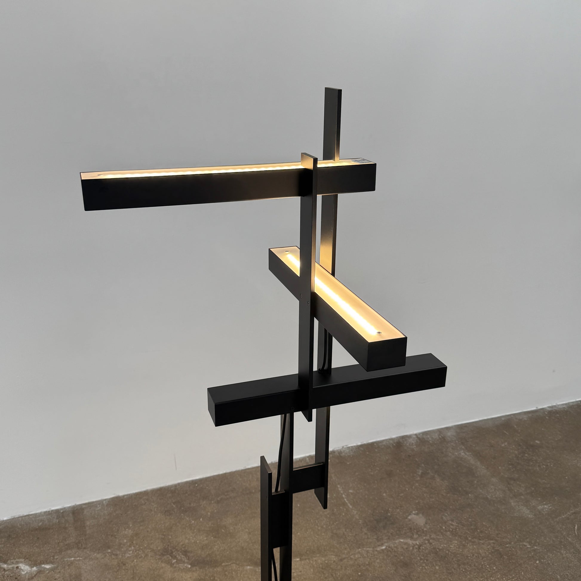 The Oluce LAS 376 LED Floor Lamp by Oluce features intersecting black rectangular panels on a flat base, with a visible power cord. Its sculptural design makes it a striking contemporary statement piece for any space.