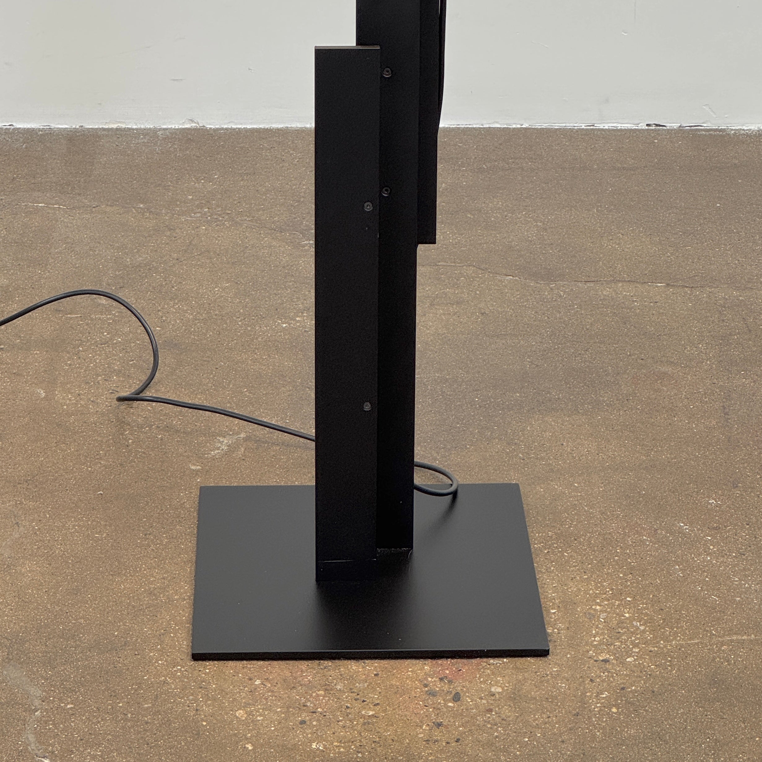 The Oluce LAS 376 LED Floor Lamp by Oluce features intersecting black rectangular panels on a flat base, with a visible power cord. Its sculptural design makes it a striking contemporary statement piece for any space.