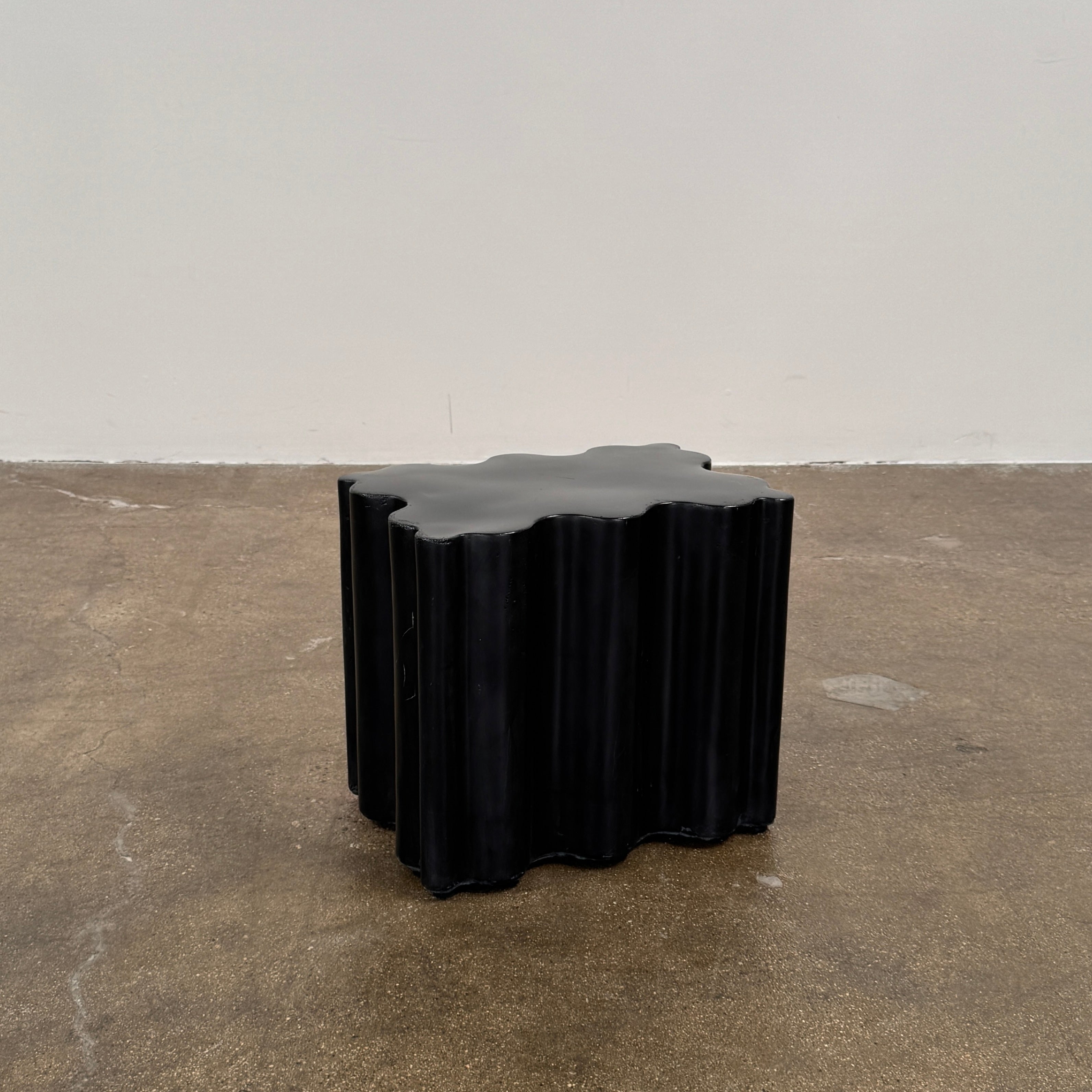 The Patrick Cain Cloud Table Black Concrete, a black wavy-edged cube by Patrick Cain, sits on a concrete floor against a plain white wall.