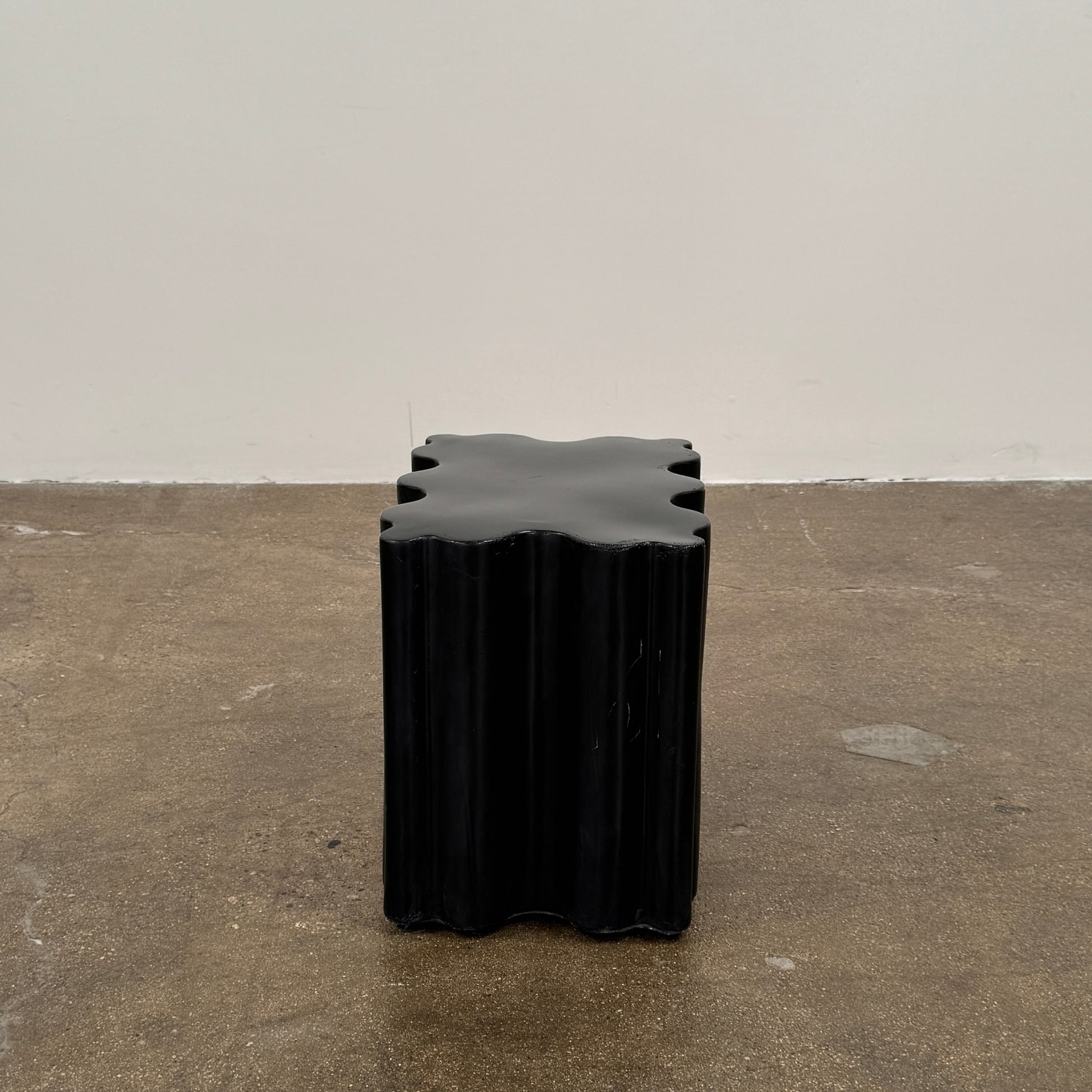 The Patrick Cain Cloud Table Black Concrete, a black wavy-edged cube by Patrick Cain, sits on a concrete floor against a plain white wall.