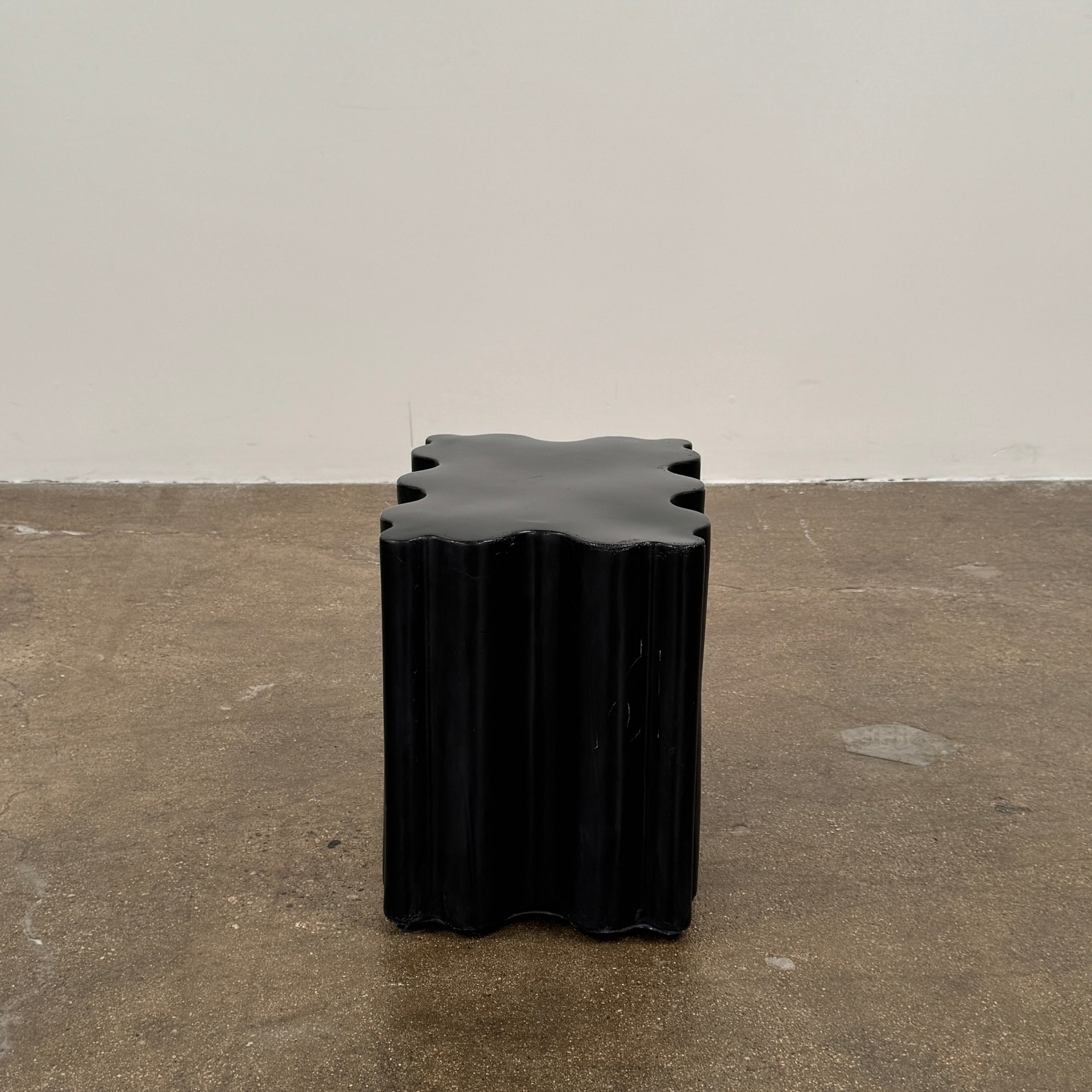 The Patrick Cain Cloud Table Black Concrete, a black wavy-edged cube by Patrick Cain, sits on a concrete floor against a plain white wall.