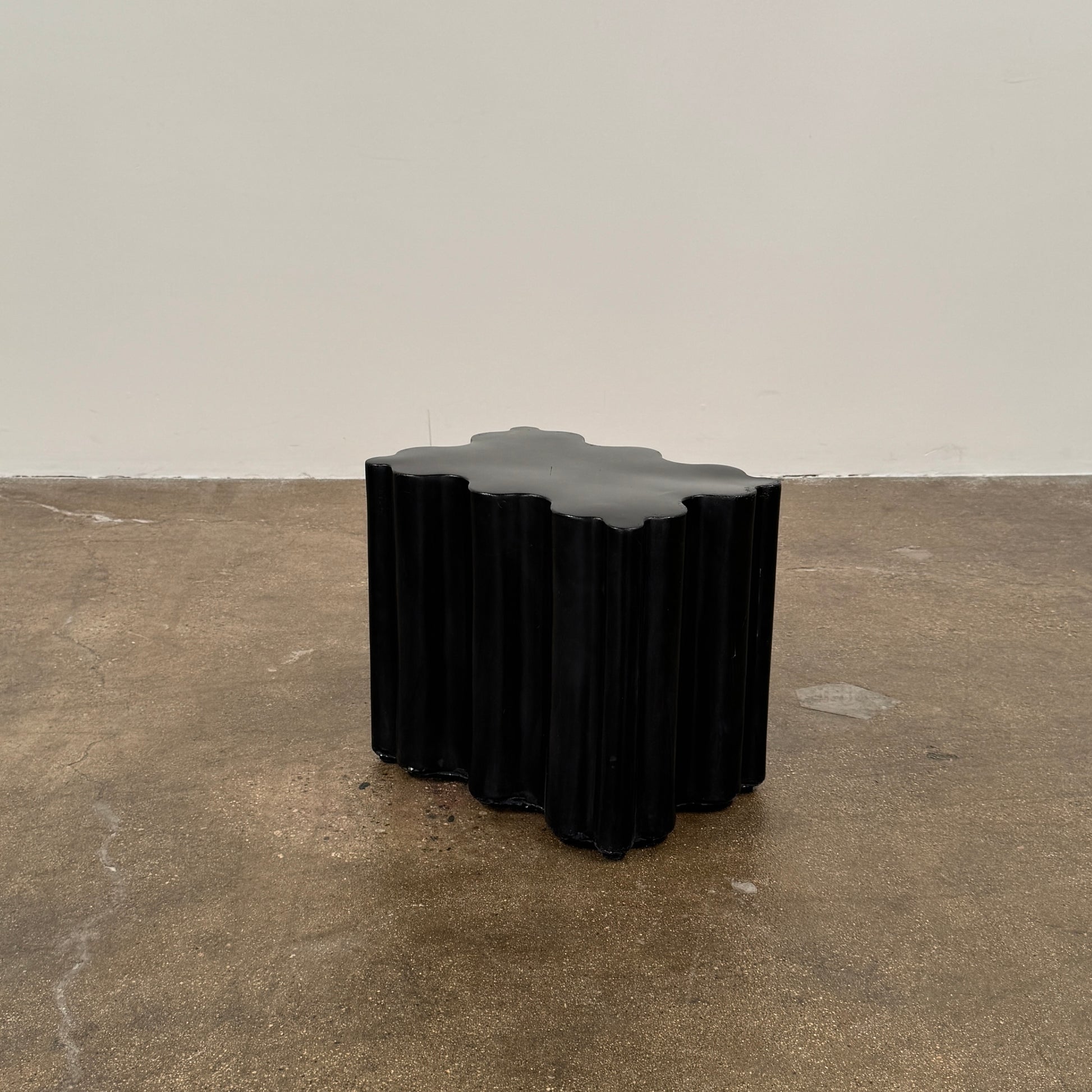 The Patrick Cain Cloud Table Black Concrete, a black wavy-edged cube by Patrick Cain, sits on a concrete floor against a plain white wall.