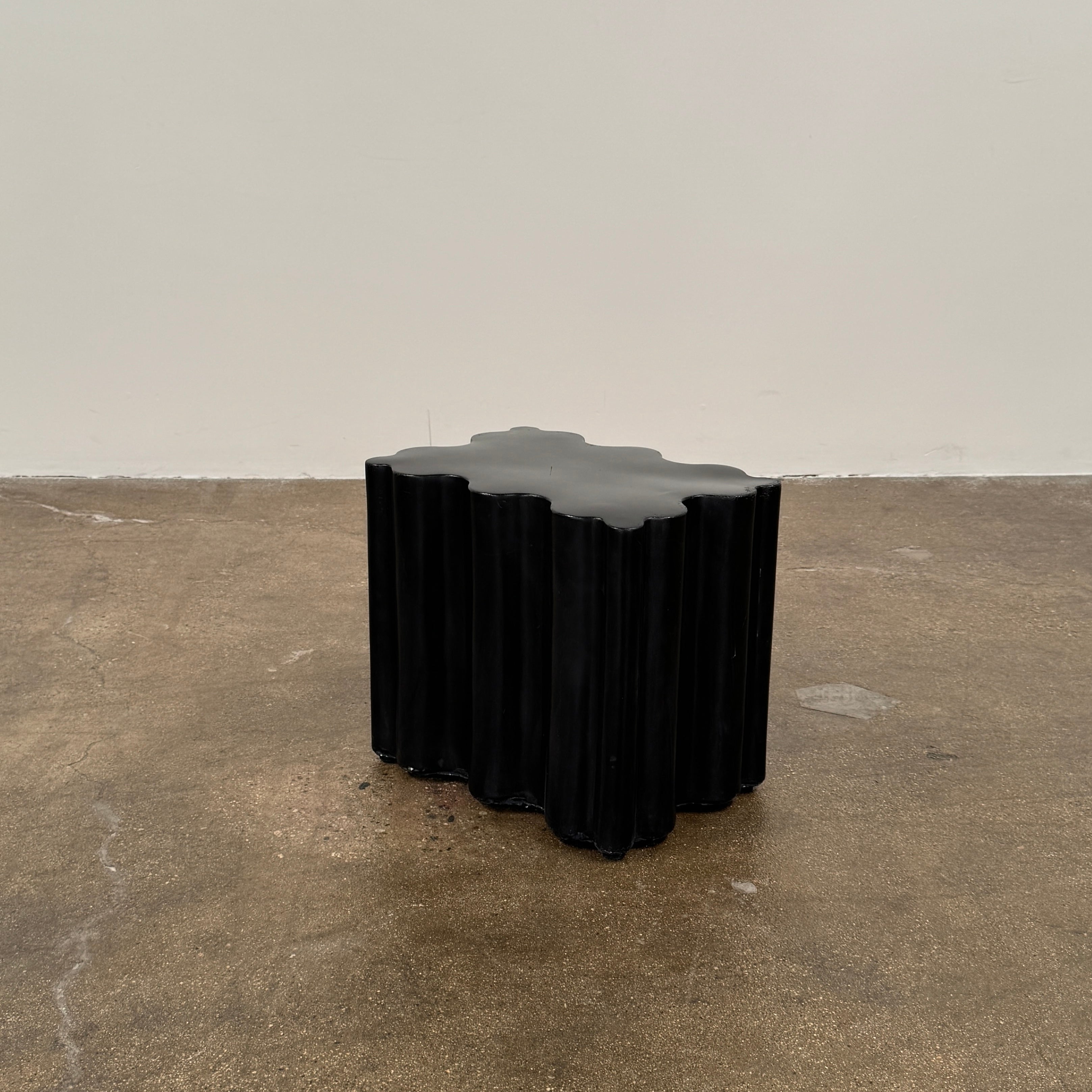 The Patrick Cain Cloud Table Black Concrete, a black wavy-edged cube by Patrick Cain, sits on a concrete floor against a plain white wall.