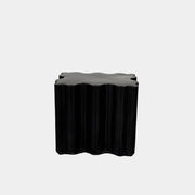 The Patrick Cain Cloud Table Black Concrete, a black wavy-edged cube by Patrick Cain, sits on a concrete floor against a plain white wall.