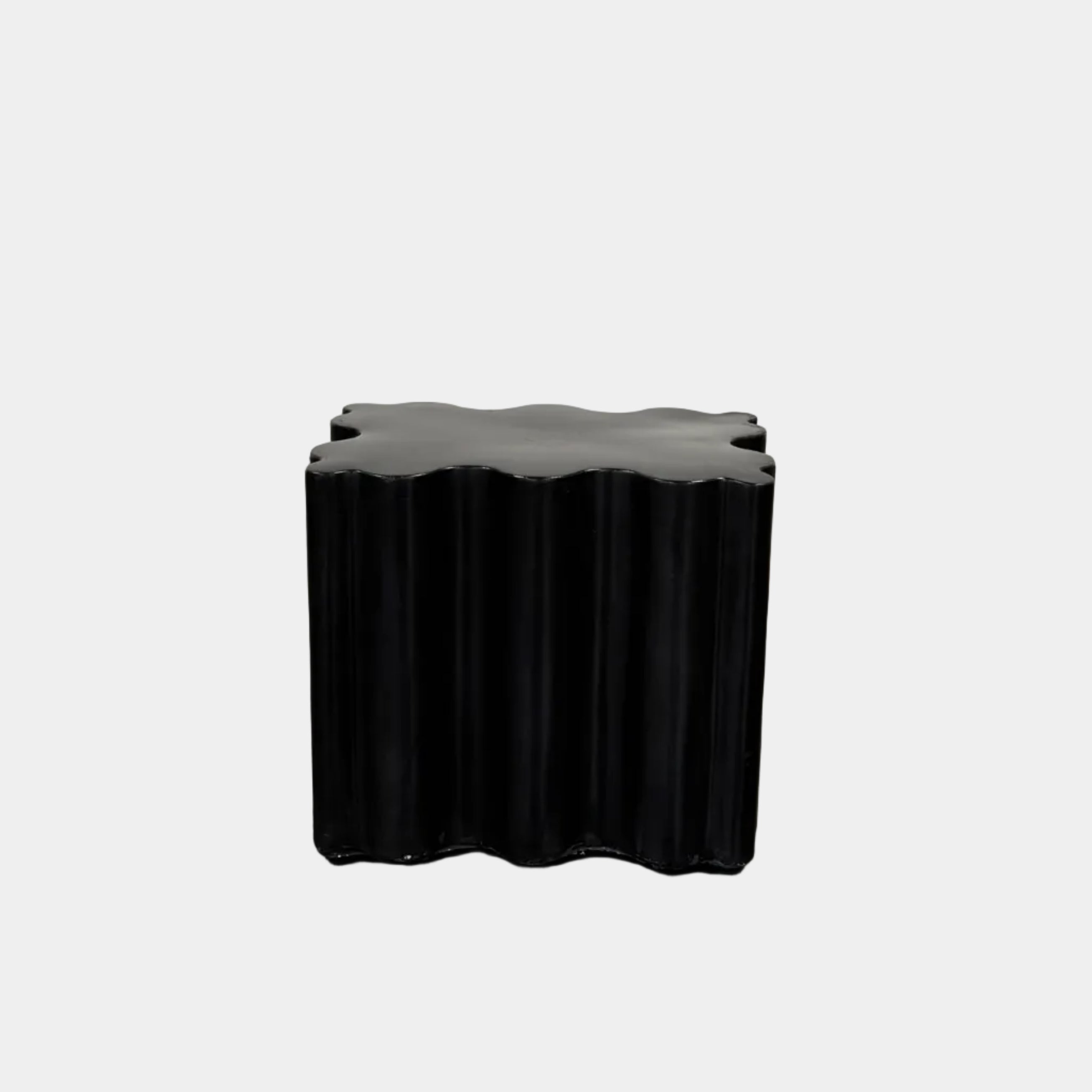 The Patrick Cain Cloud Table Black Concrete, a black wavy-edged cube by Patrick Cain, sits on a concrete floor against a plain white wall.