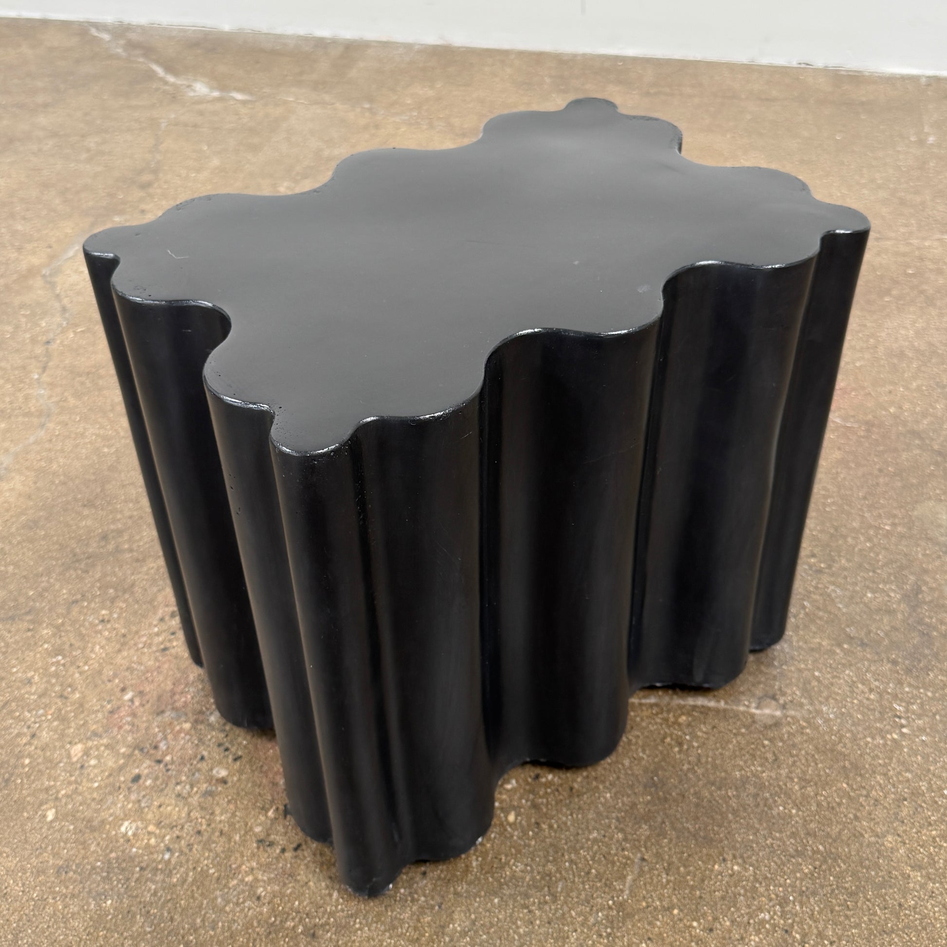 The Patrick Cain Cloud Table Black Concrete, a black wavy-edged cube by Patrick Cain, sits on a concrete floor against a plain white wall.