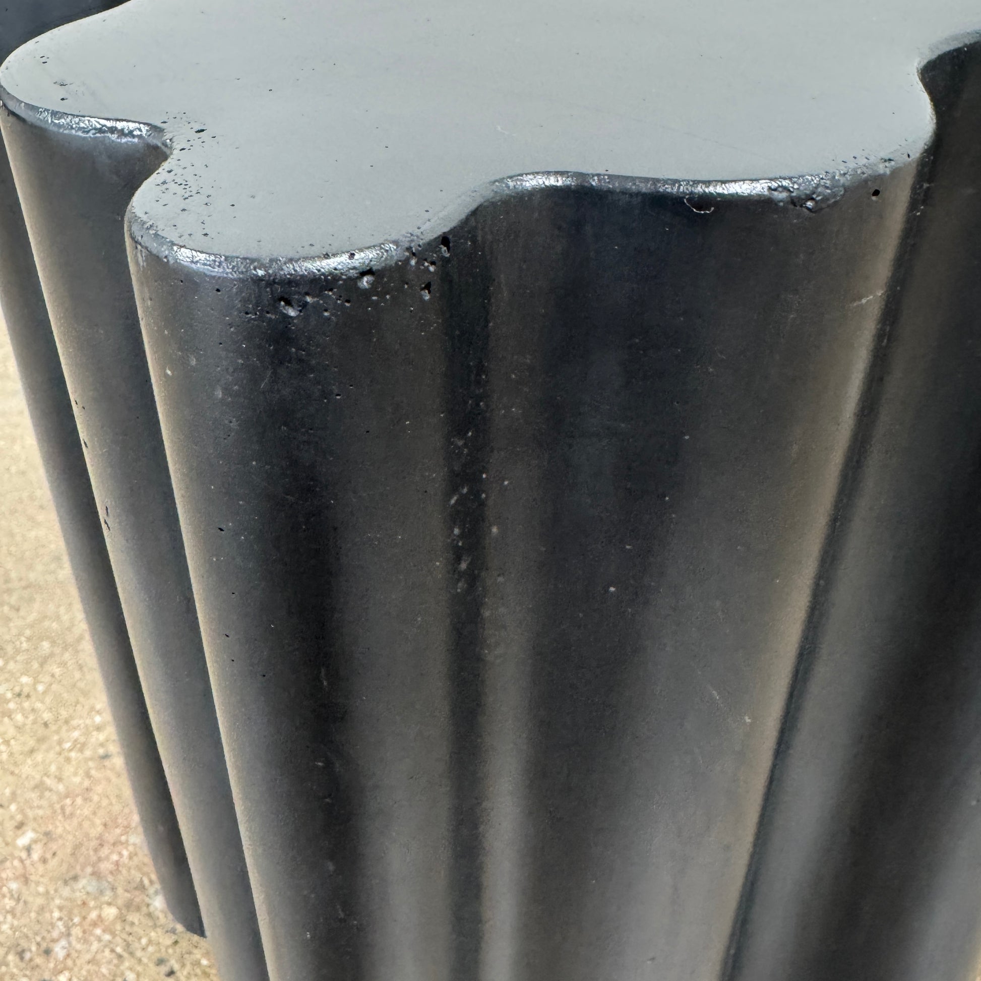 The Patrick Cain Cloud Table Black Concrete, a black wavy-edged cube by Patrick Cain, sits on a concrete floor against a plain white wall.