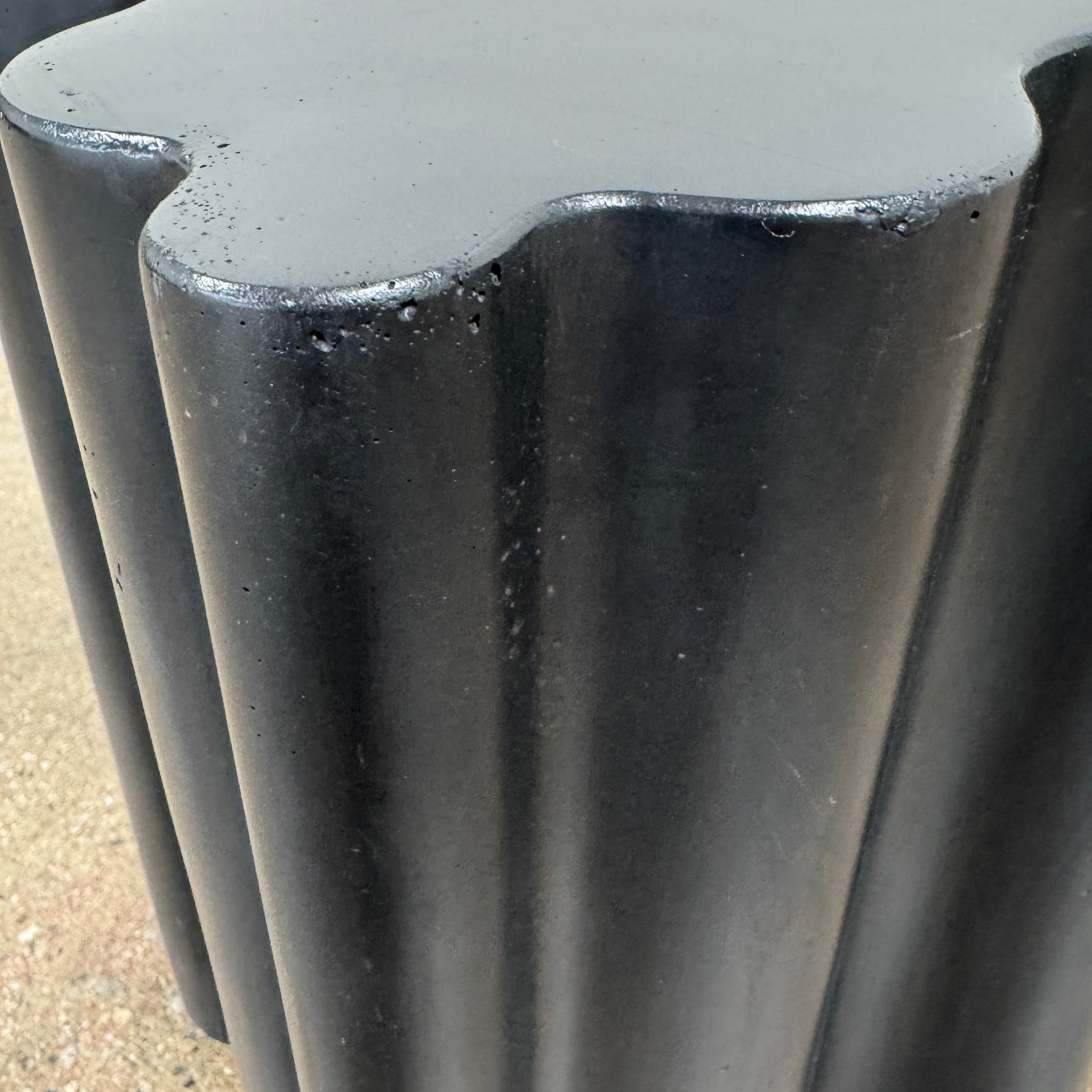 The Patrick Cain Cloud Table Black Concrete, a black wavy-edged cube by Patrick Cain, sits on a concrete floor against a plain white wall.