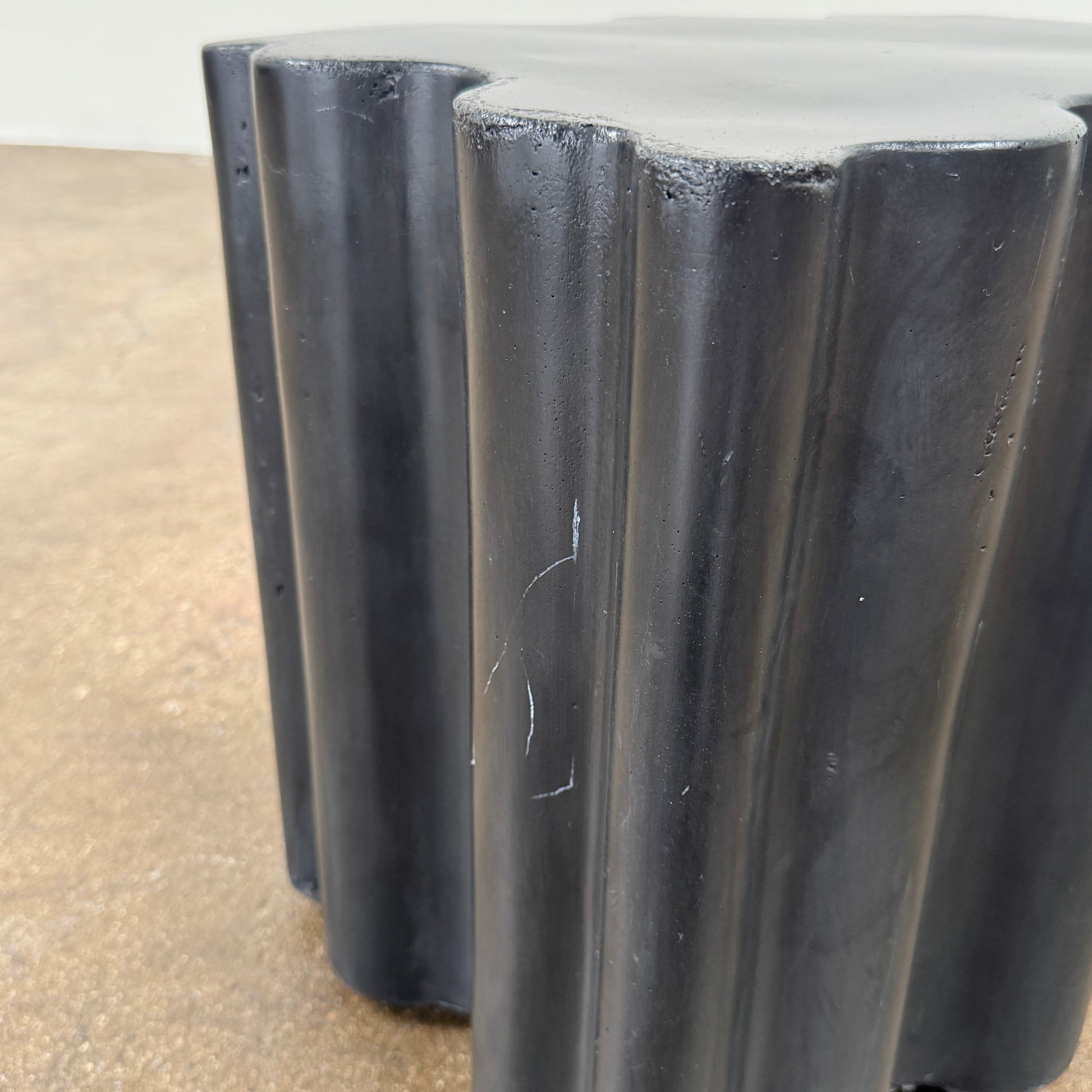 The Patrick Cain Cloud Table Black Concrete, a black wavy-edged cube by Patrick Cain, sits on a concrete floor against a plain white wall.