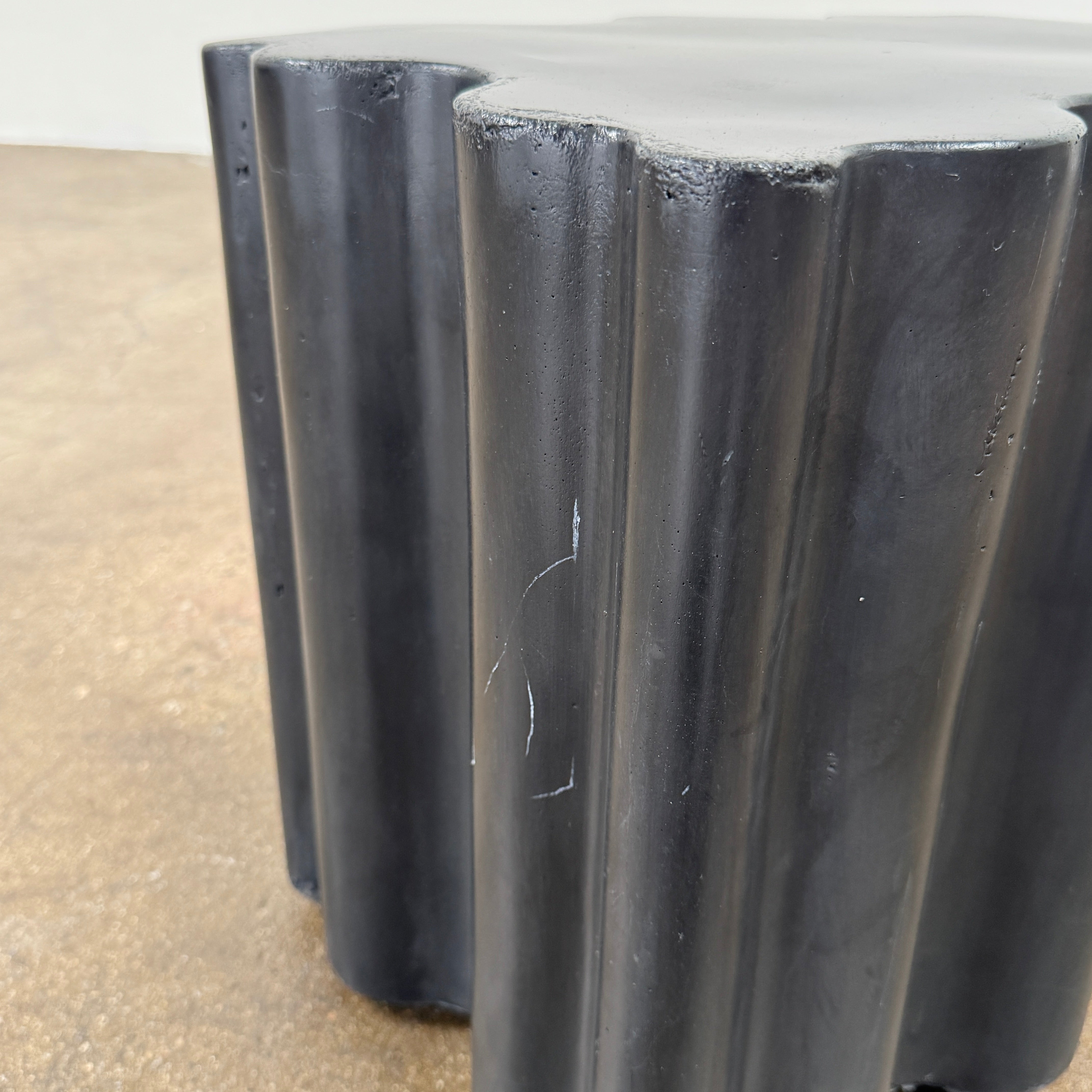 The Patrick Cain Cloud Table Black Concrete, a black wavy-edged cube by Patrick Cain, sits on a concrete floor against a plain white wall.