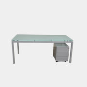 The DWR Tavolo Glass Desk with Chest of drawers by DWR features a sleek glass top, metal legs, and a mobile chest of drawers beneath—perfect for a contemporary office setup.