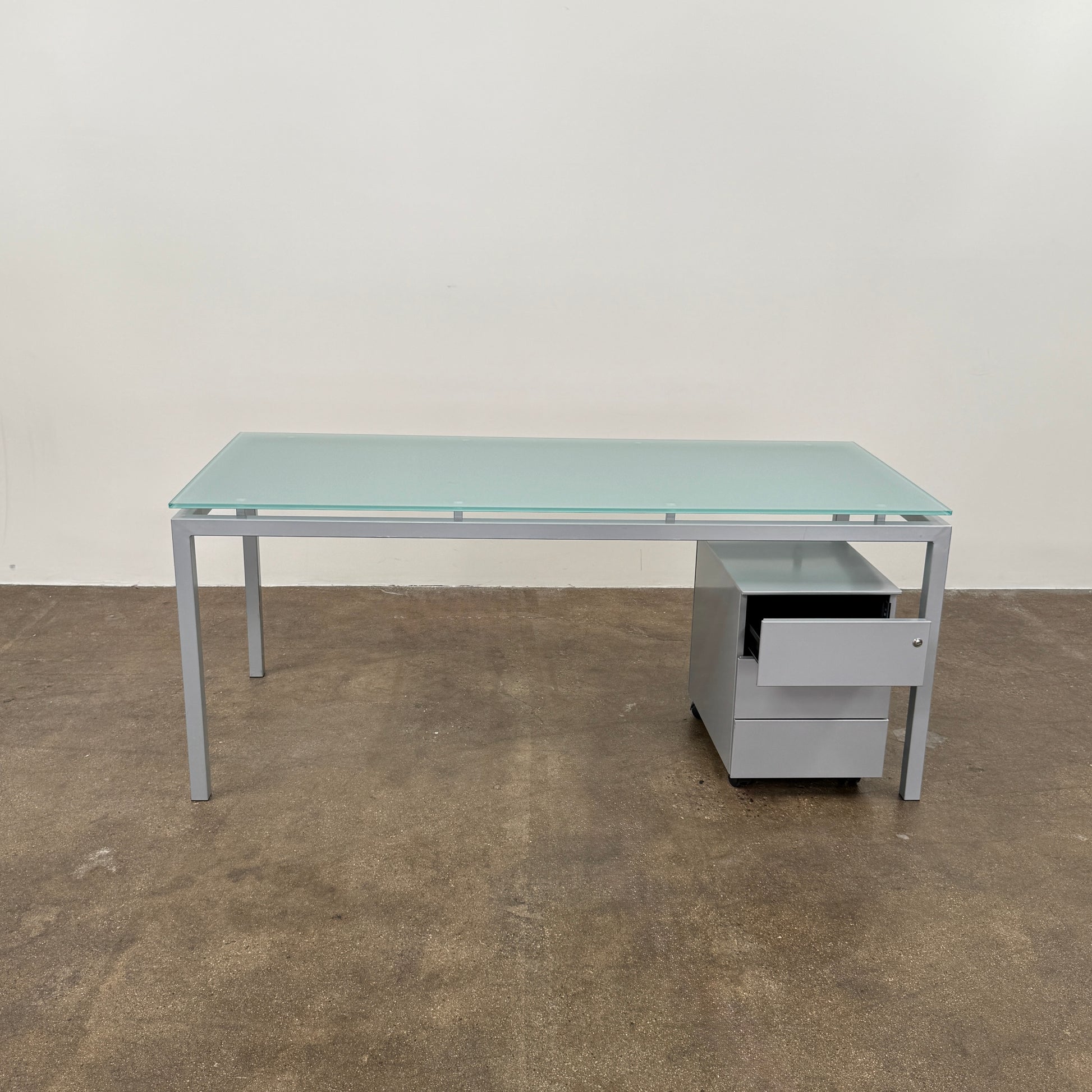 The DWR Tavolo Glass Desk with Chest of drawers by DWR features a sleek glass top, metal legs, and a mobile chest of drawers beneath—perfect for a contemporary office setup.