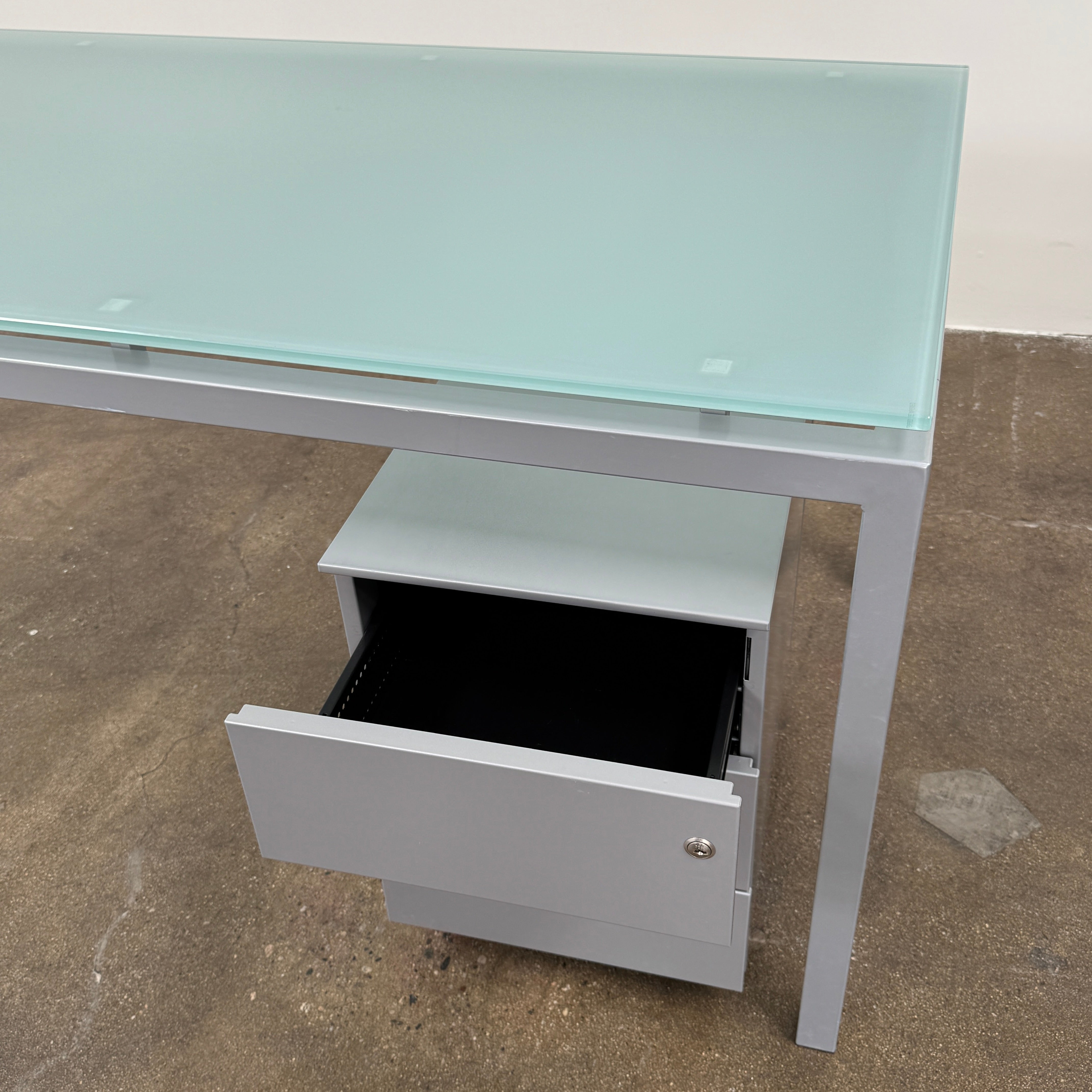 The DWR Tavolo Glass Desk with Chest of drawers by DWR features a sleek glass top, metal legs, and a mobile chest of drawers beneath—perfect for a contemporary office setup.