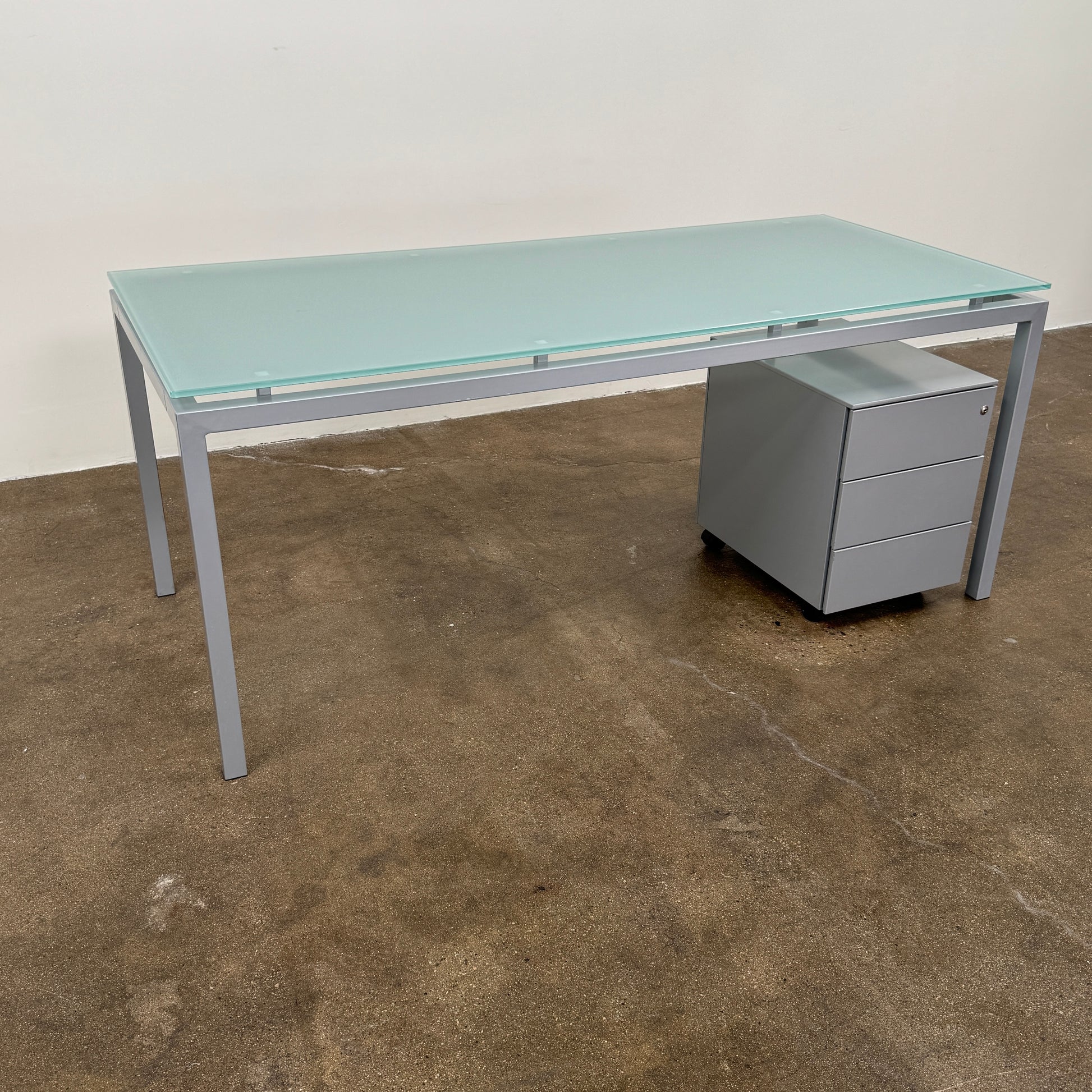 The DWR Tavolo Glass Desk with Chest of drawers by DWR features a sleek glass top, metal legs, and a mobile chest of drawers beneath—perfect for a contemporary office setup.