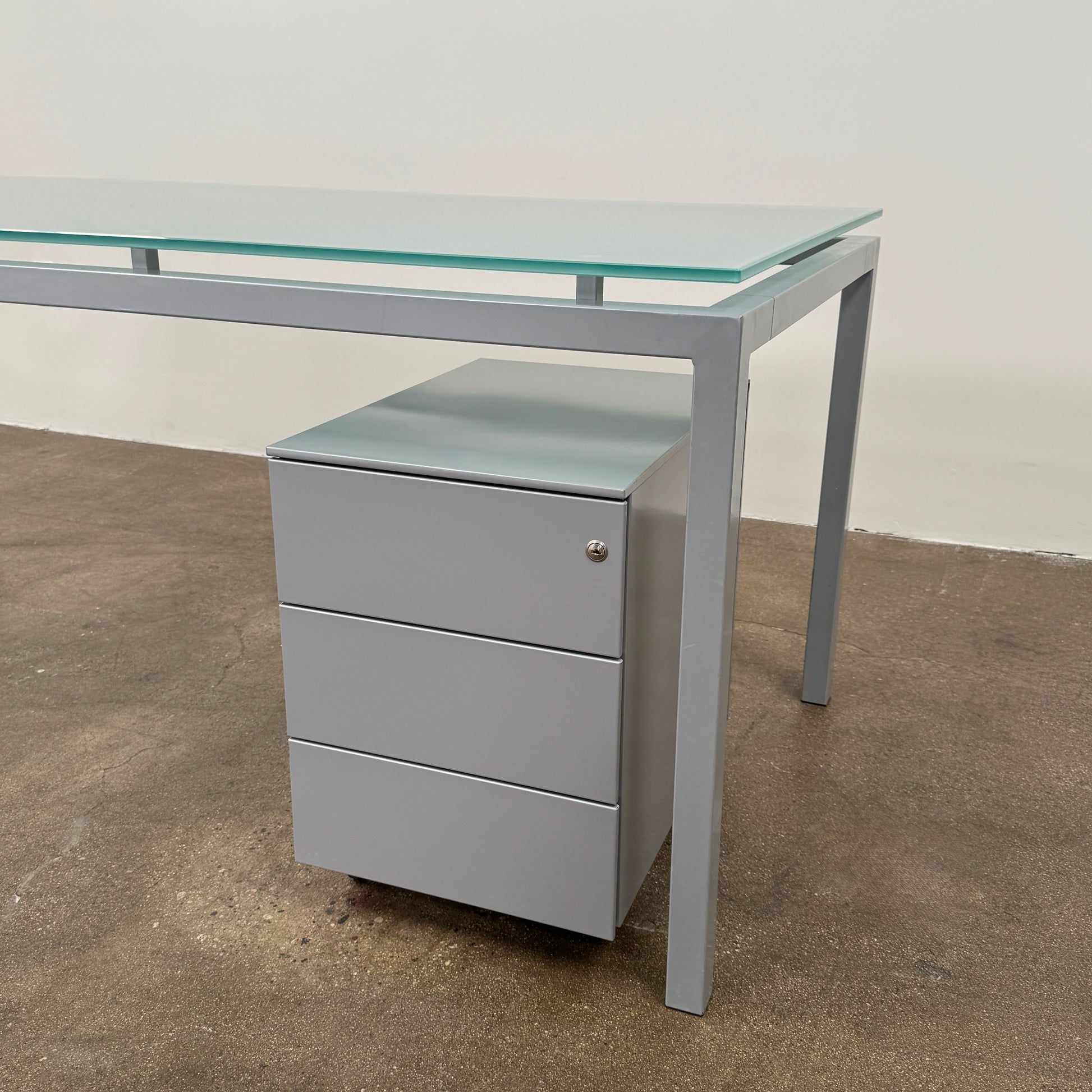 The DWR Tavolo Glass Desk with Chest of drawers by DWR features a sleek glass top, metal legs, and a mobile chest of drawers beneath—perfect for a contemporary office setup.