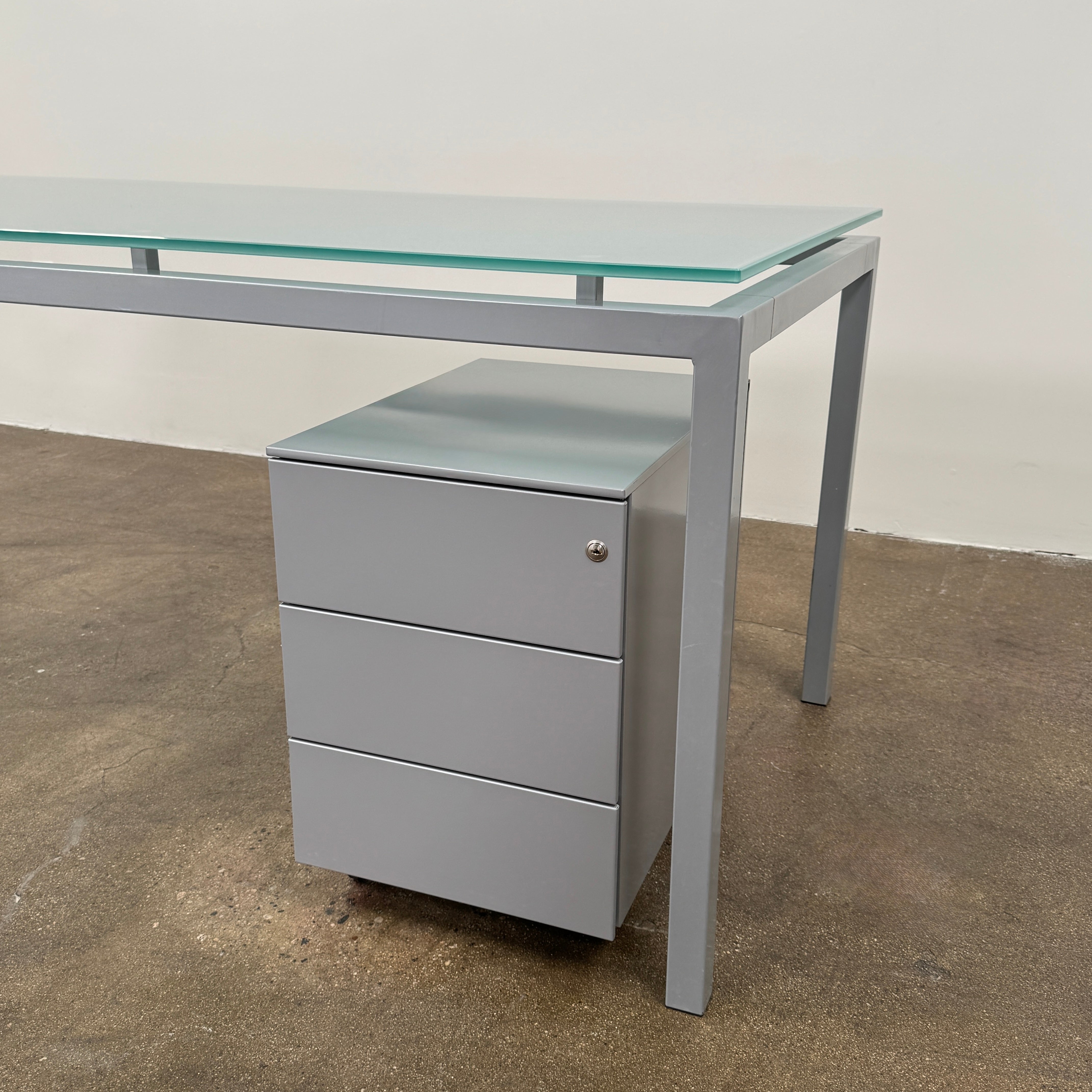 The DWR Tavolo Glass Desk with Chest of drawers by DWR features a sleek glass top, metal legs, and a mobile chest of drawers beneath—perfect for a contemporary office setup.