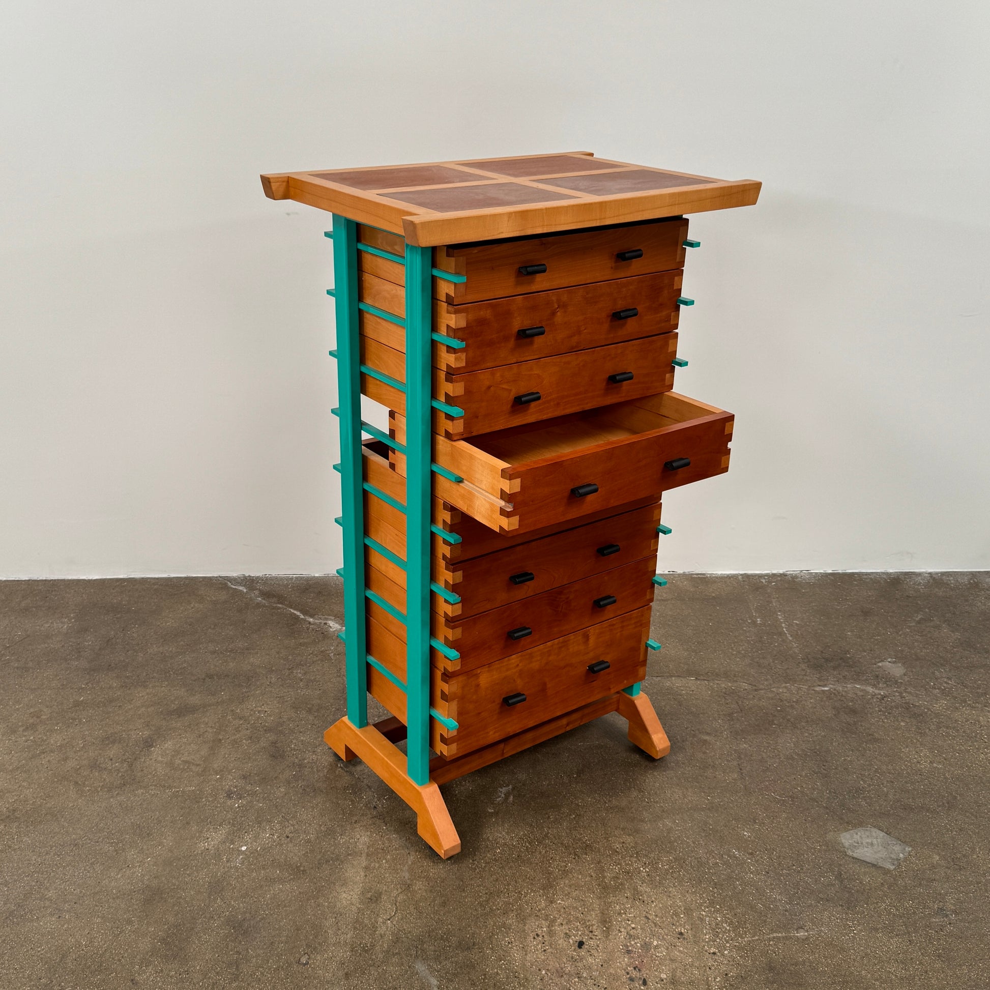 The Joseph Schwarte Tall Chest of Drawers by Artist features nine drawers, teal vertical side supports, black rectangular pulls, and a flat top—postmodern-inspired and shown against a plain background.