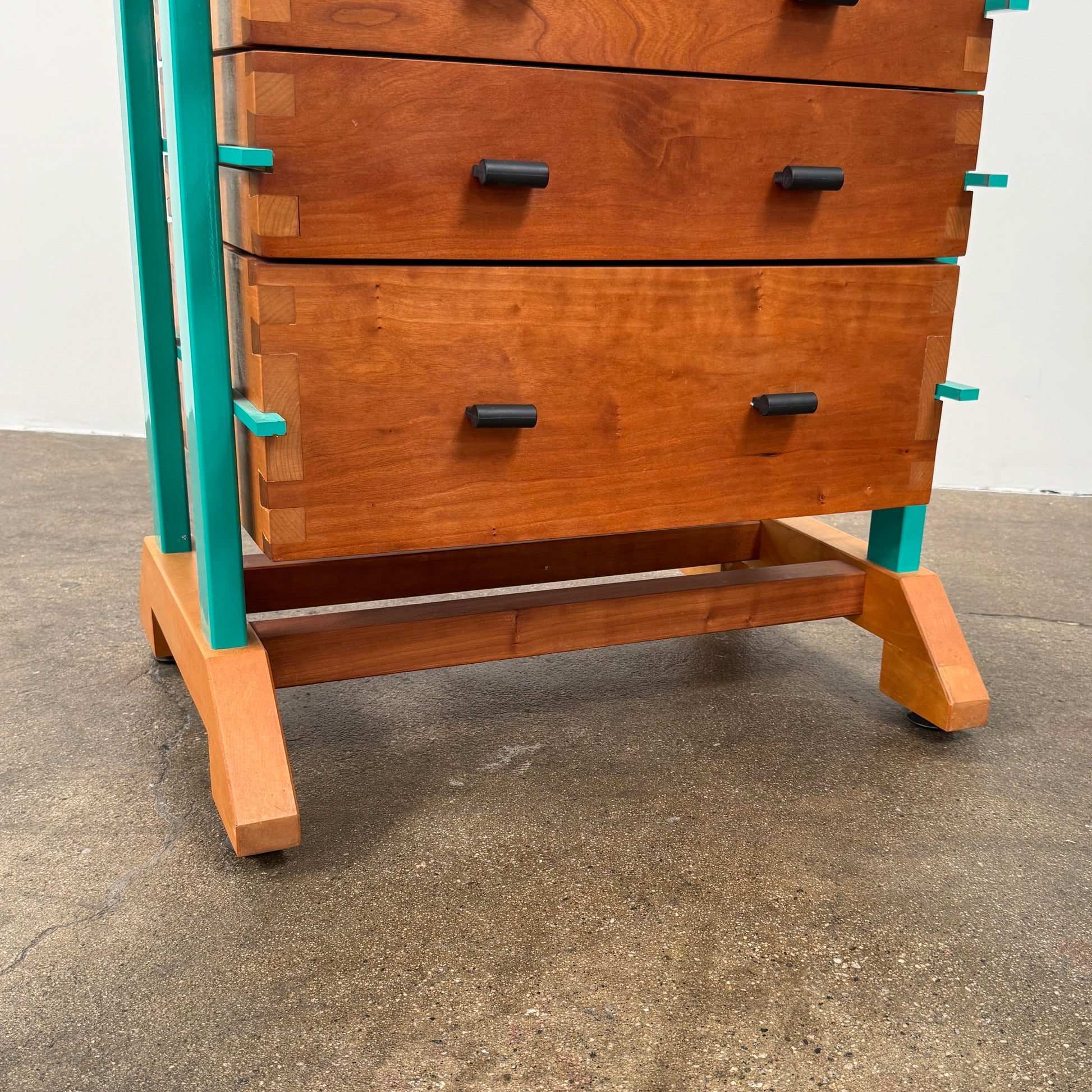 The Joseph Schwarte Tall Chest of Drawers by Artist features nine drawers, teal vertical side supports, black rectangular pulls, and a flat top—postmodern-inspired and shown against a plain background.