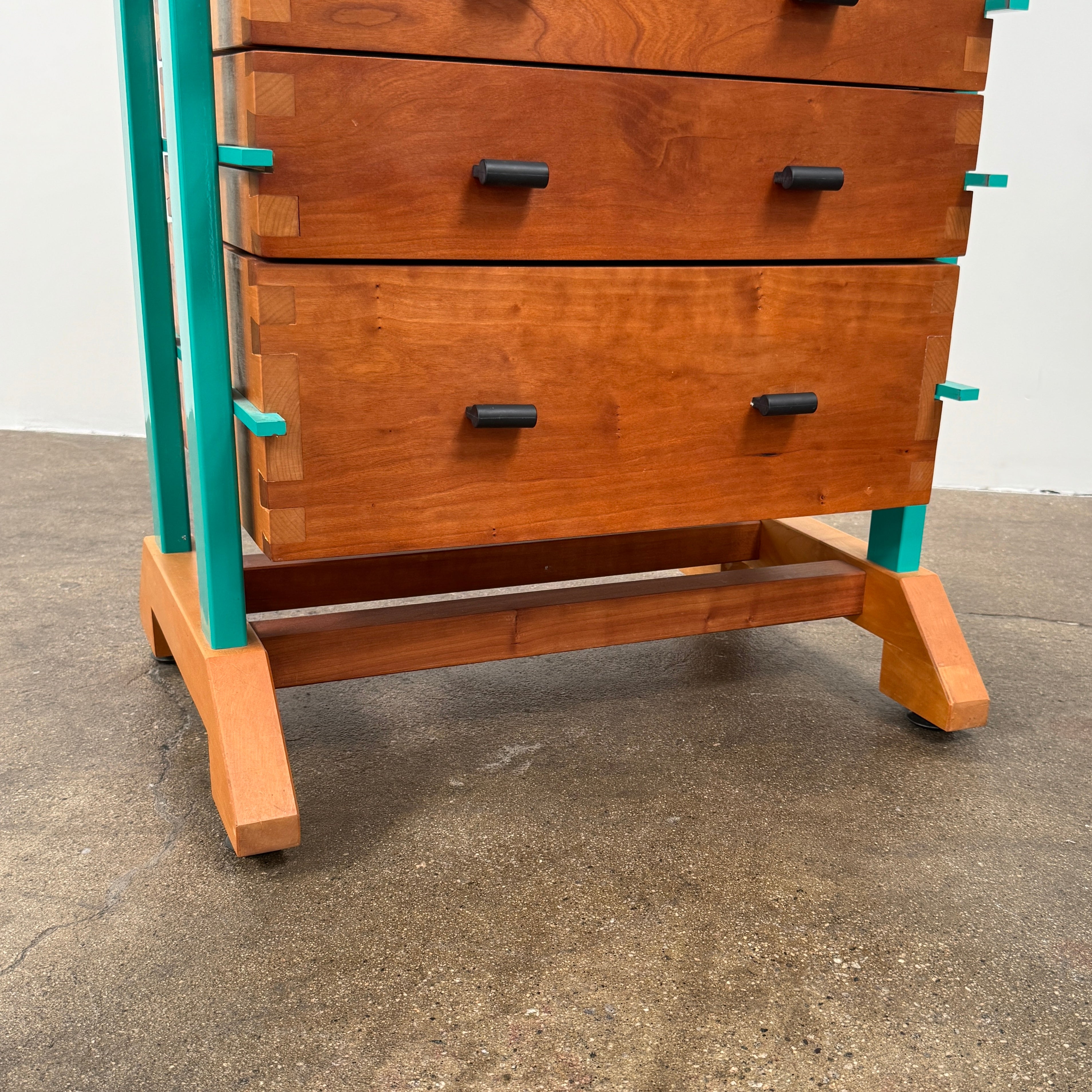 The Joseph Schwarte Tall Chest of Drawers by Artist features nine drawers, teal vertical side supports, black rectangular pulls, and a flat top—postmodern-inspired and shown against a plain background.