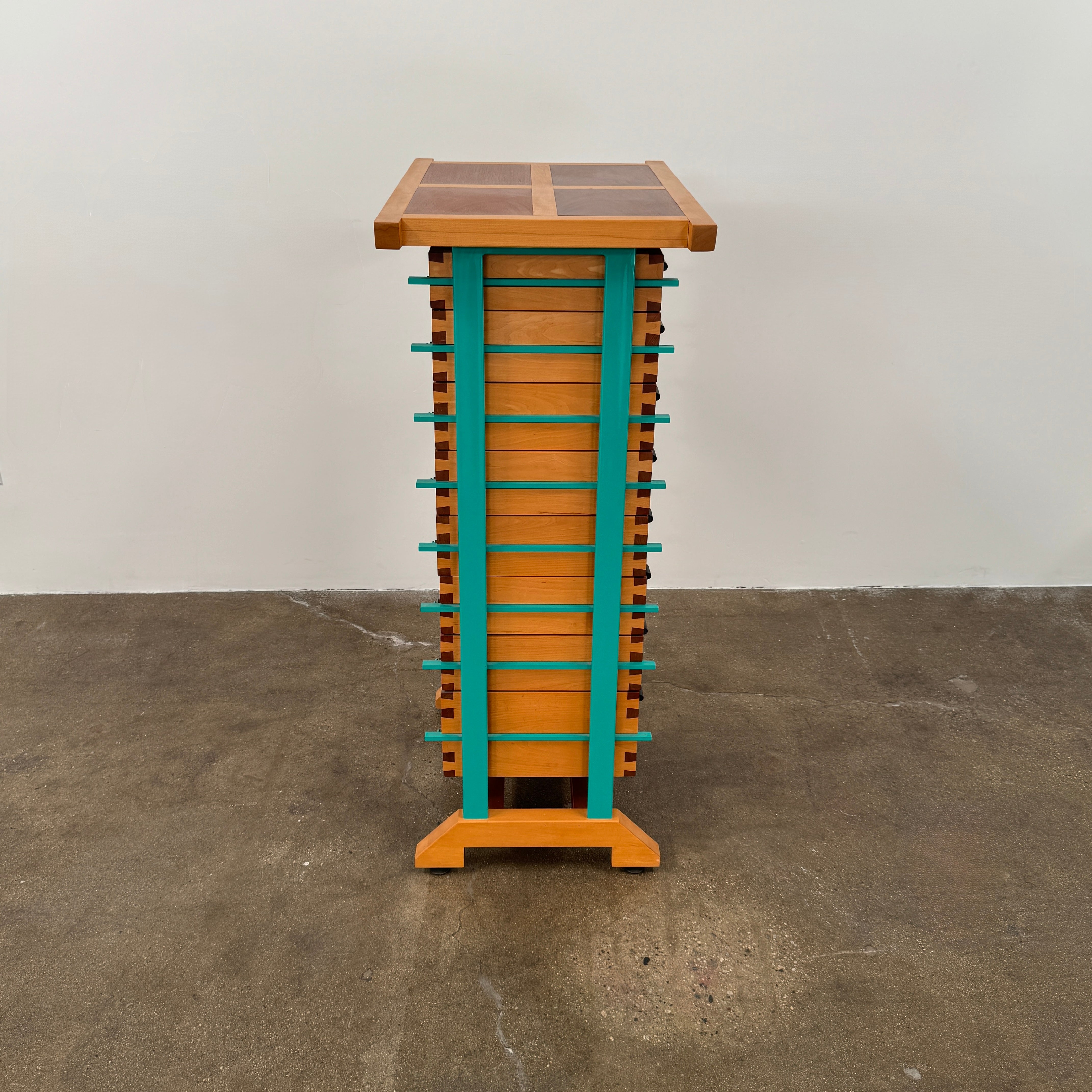 The Joseph Schwarte Tall Chest of Drawers by Artist features nine drawers, teal vertical side supports, black rectangular pulls, and a flat top—postmodern-inspired and shown against a plain background.
