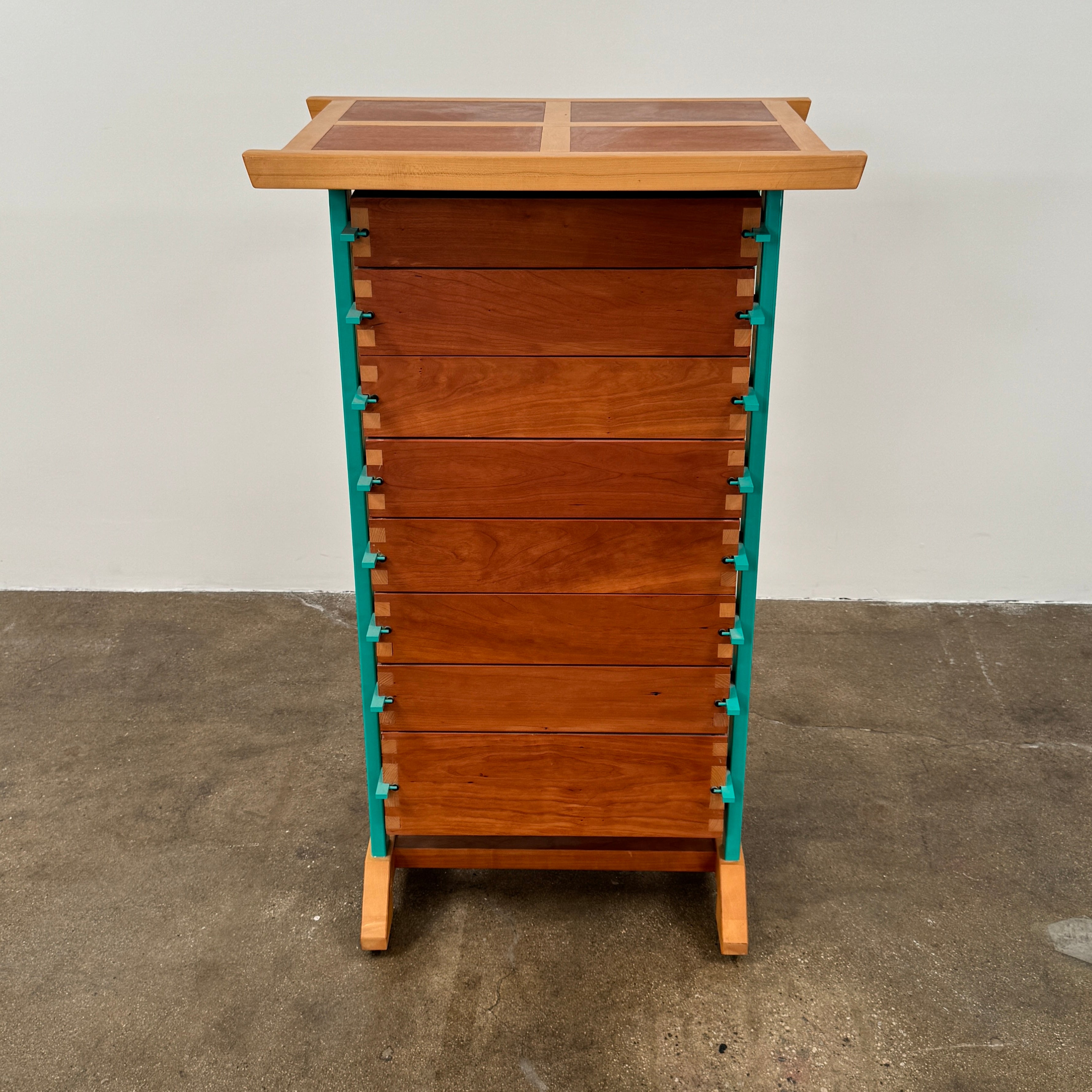 The Joseph Schwarte Tall Chest of Drawers by Artist features nine drawers, teal vertical side supports, black rectangular pulls, and a flat top—postmodern-inspired and shown against a plain background.