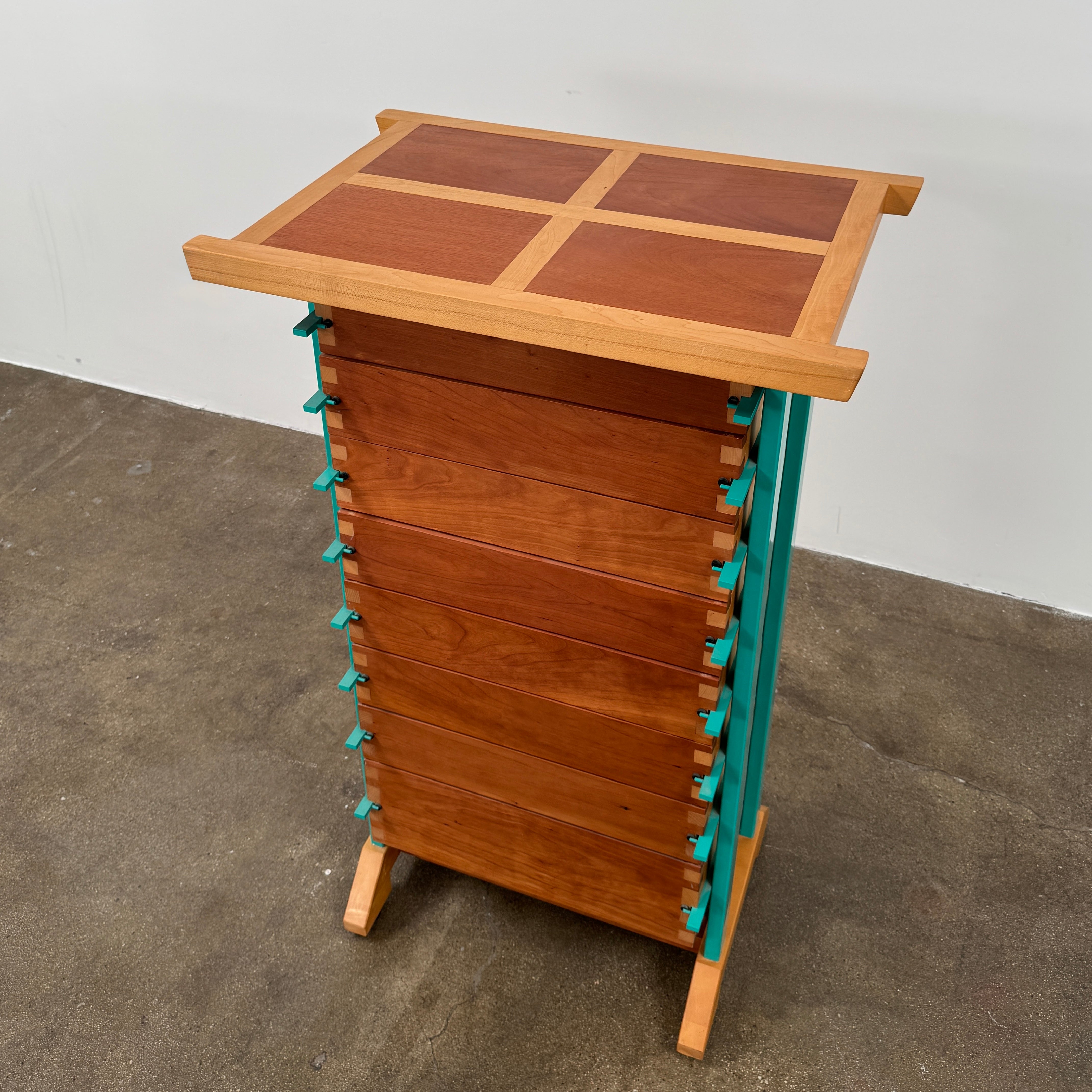 The Joseph Schwarte Tall Chest of Drawers by Artist features nine drawers, teal vertical side supports, black rectangular pulls, and a flat top—postmodern-inspired and shown against a plain background.
