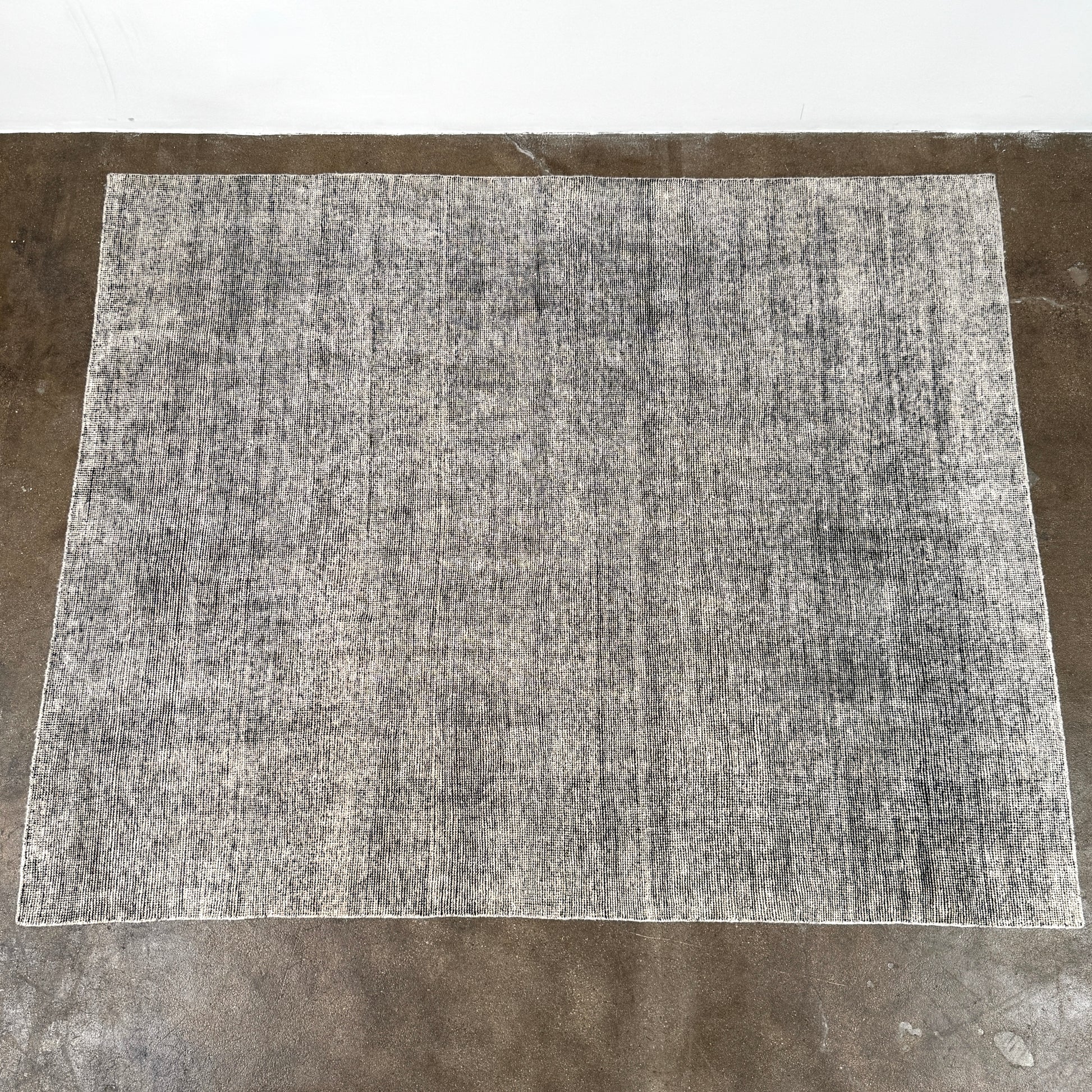 An 8'x10' rectangular DWR Luna Rug in gray with a subtle textured pattern is placed on a concrete floor against a white wall.