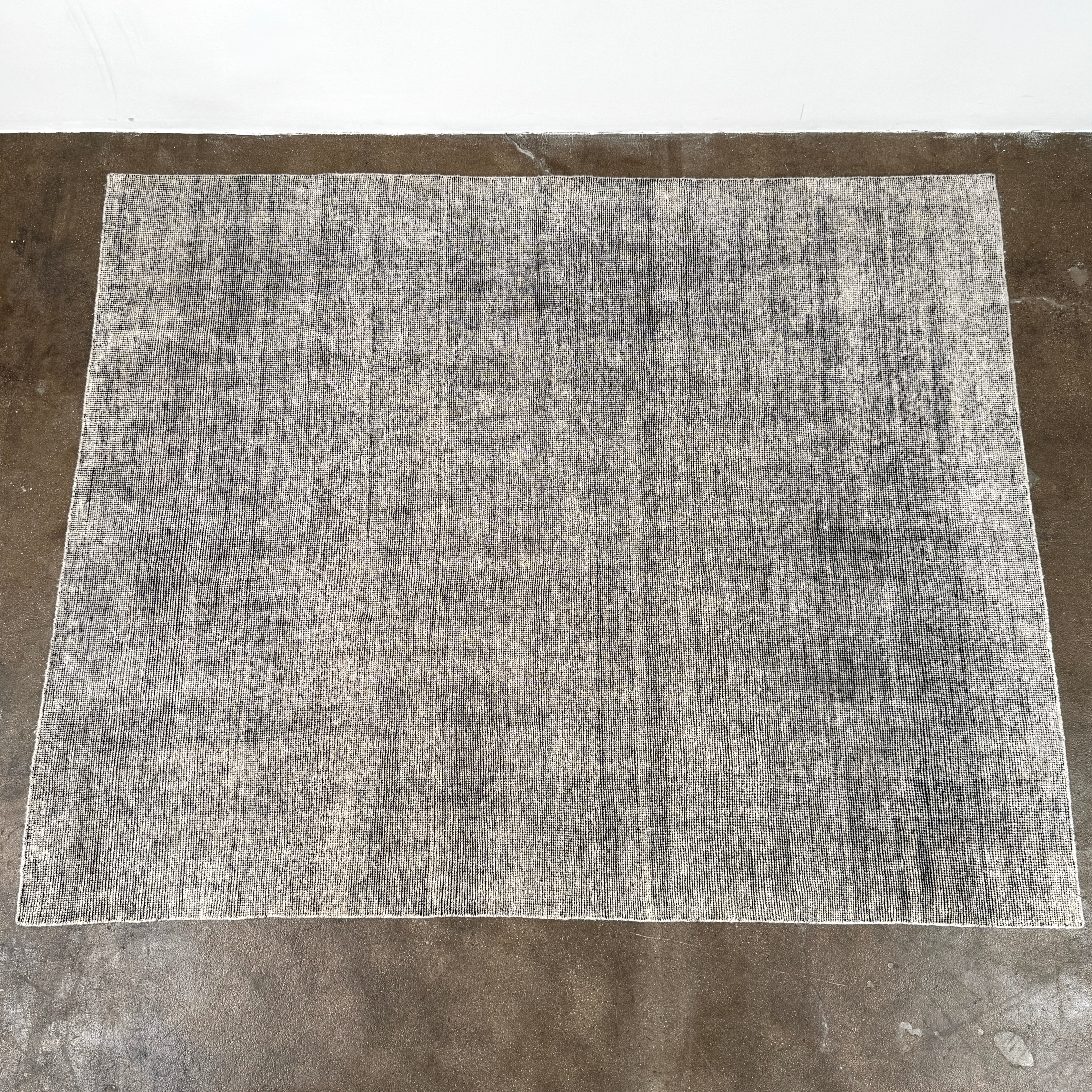 An 8'x10' rectangular DWR Luna Rug in gray with a subtle textured pattern is placed on a concrete floor against a white wall.