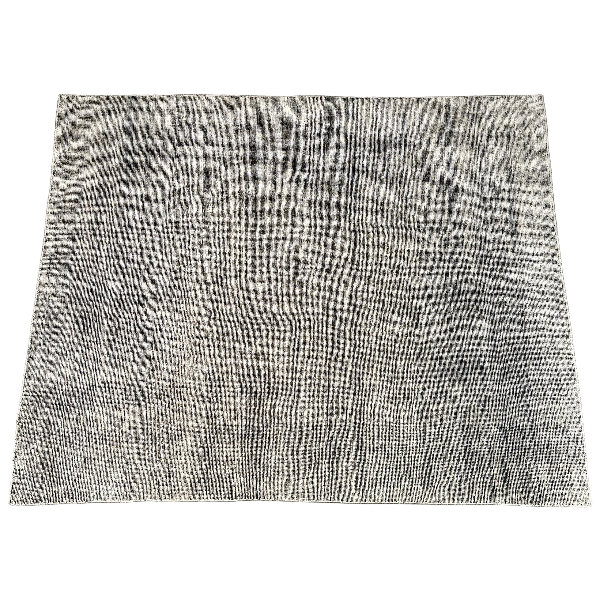 An 8'x10' rectangular DWR Luna Rug in gray with a subtle textured pattern is placed on a concrete floor against a white wall.