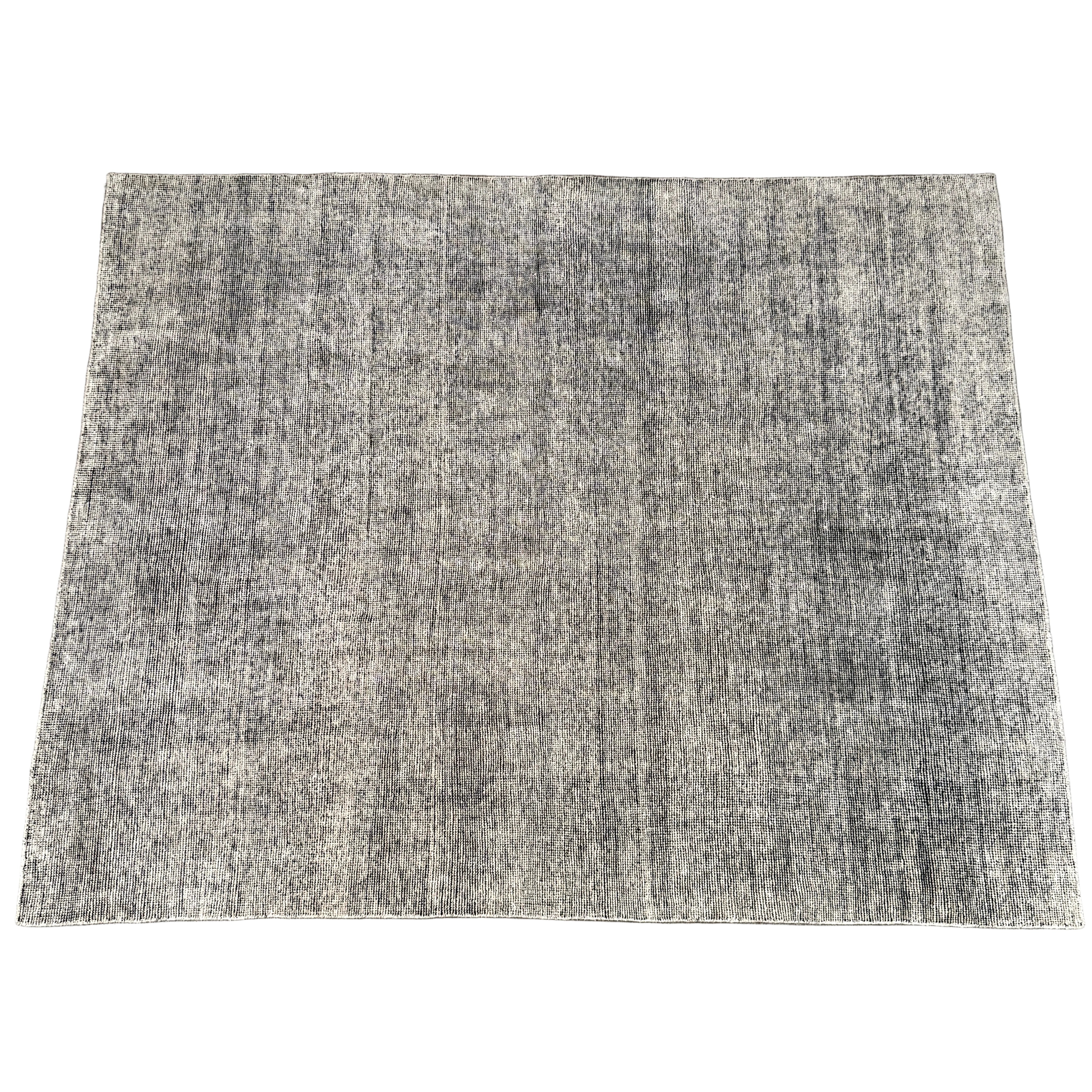An 8'x10' rectangular DWR Luna Rug in gray with a subtle textured pattern is placed on a concrete floor against a white wall.
