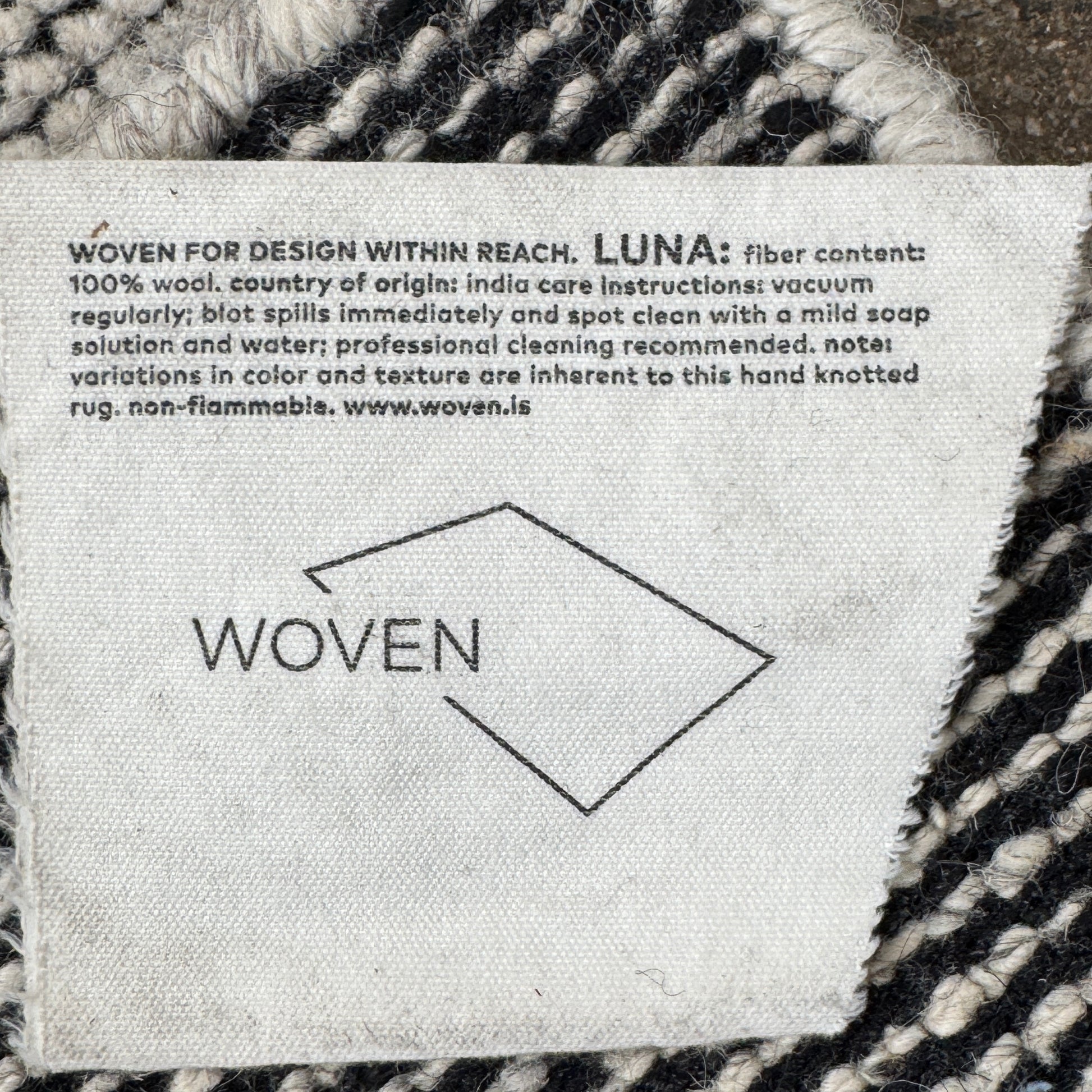 An 8'x10' rectangular DWR Luna Rug in gray with a subtle textured pattern is placed on a concrete floor against a white wall.