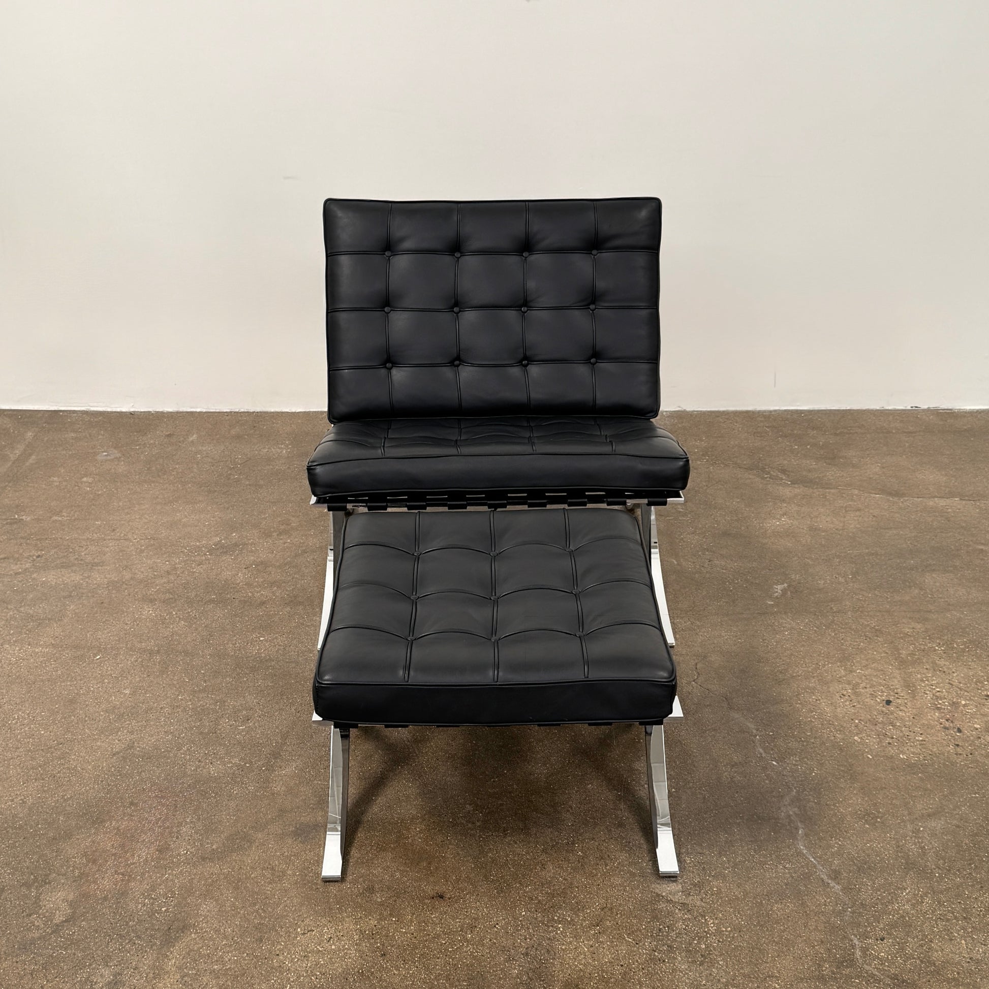 The Vintage DWR Barcelona Black Leather Chair & Ottoman by DWR features button-tufted cushions and chrome X-shaped legs, taking inspiration from the iconic Barcelona Chair, all presented against a plain white background.