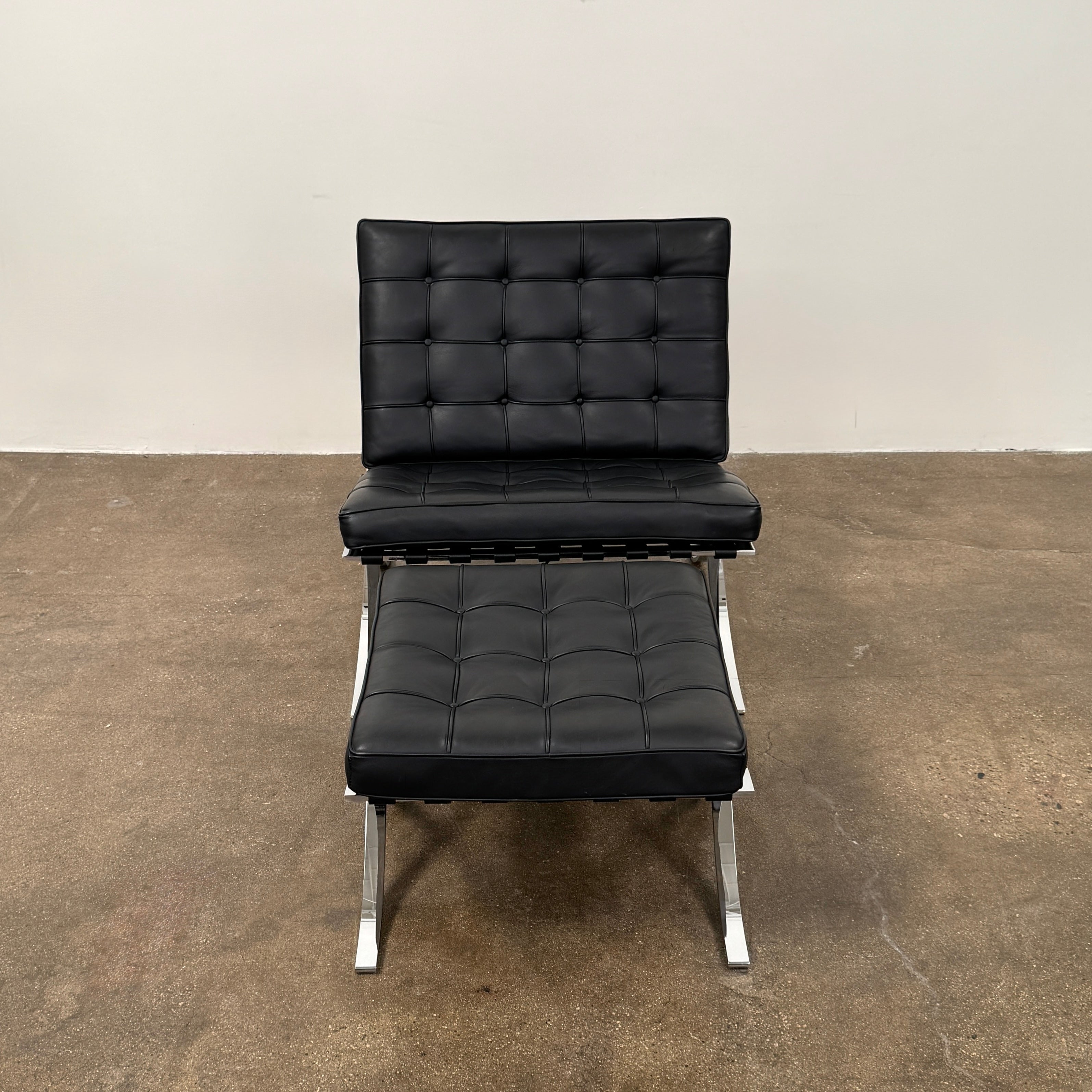 The Vintage DWR Barcelona Black Leather Chair & Ottoman by DWR features button-tufted cushions and chrome X-shaped legs, taking inspiration from the iconic Barcelona Chair, all presented against a plain white background.