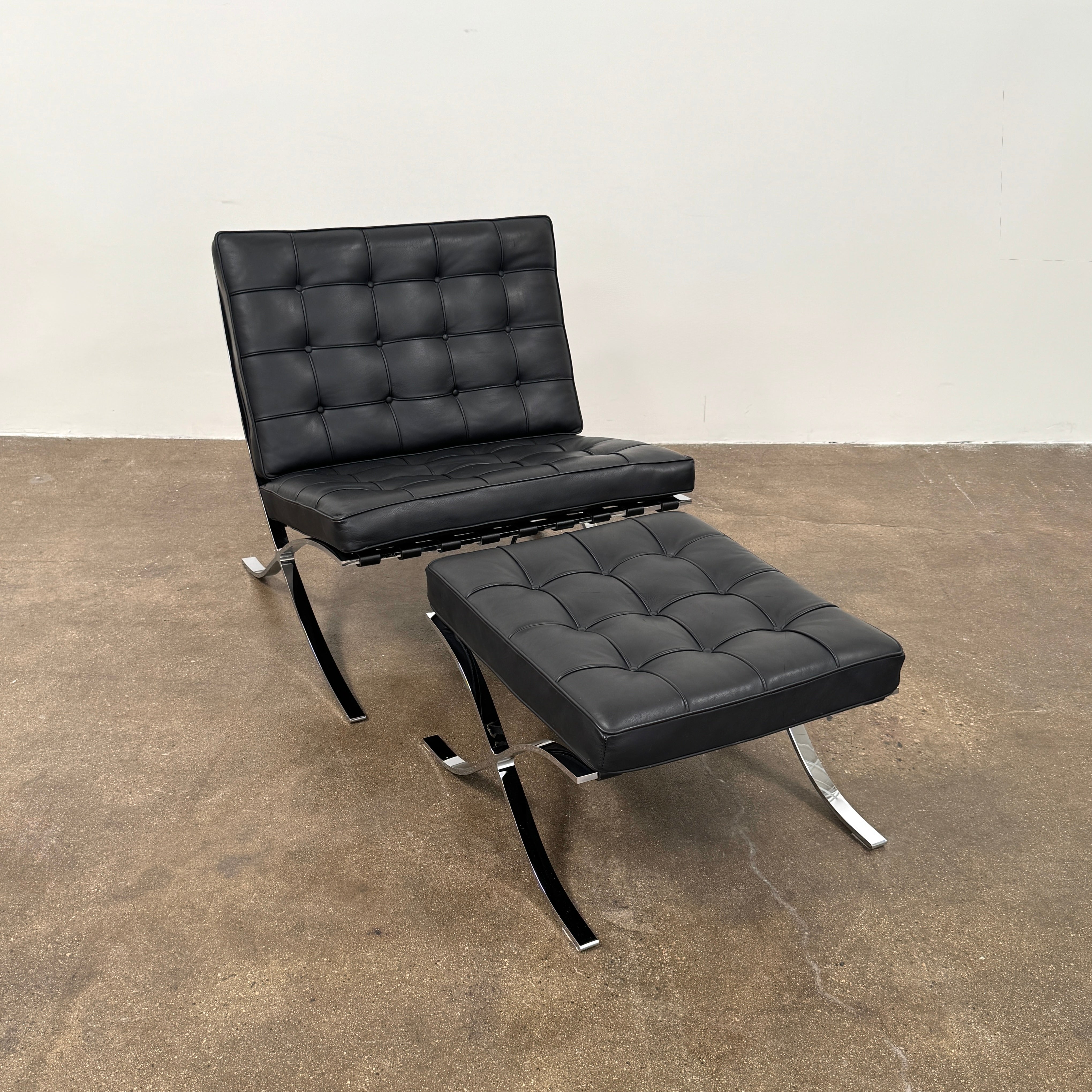 The Vintage DWR Barcelona Black Leather Chair & Ottoman by DWR features button-tufted cushions and chrome X-shaped legs, taking inspiration from the iconic Barcelona Chair, all presented against a plain white background.