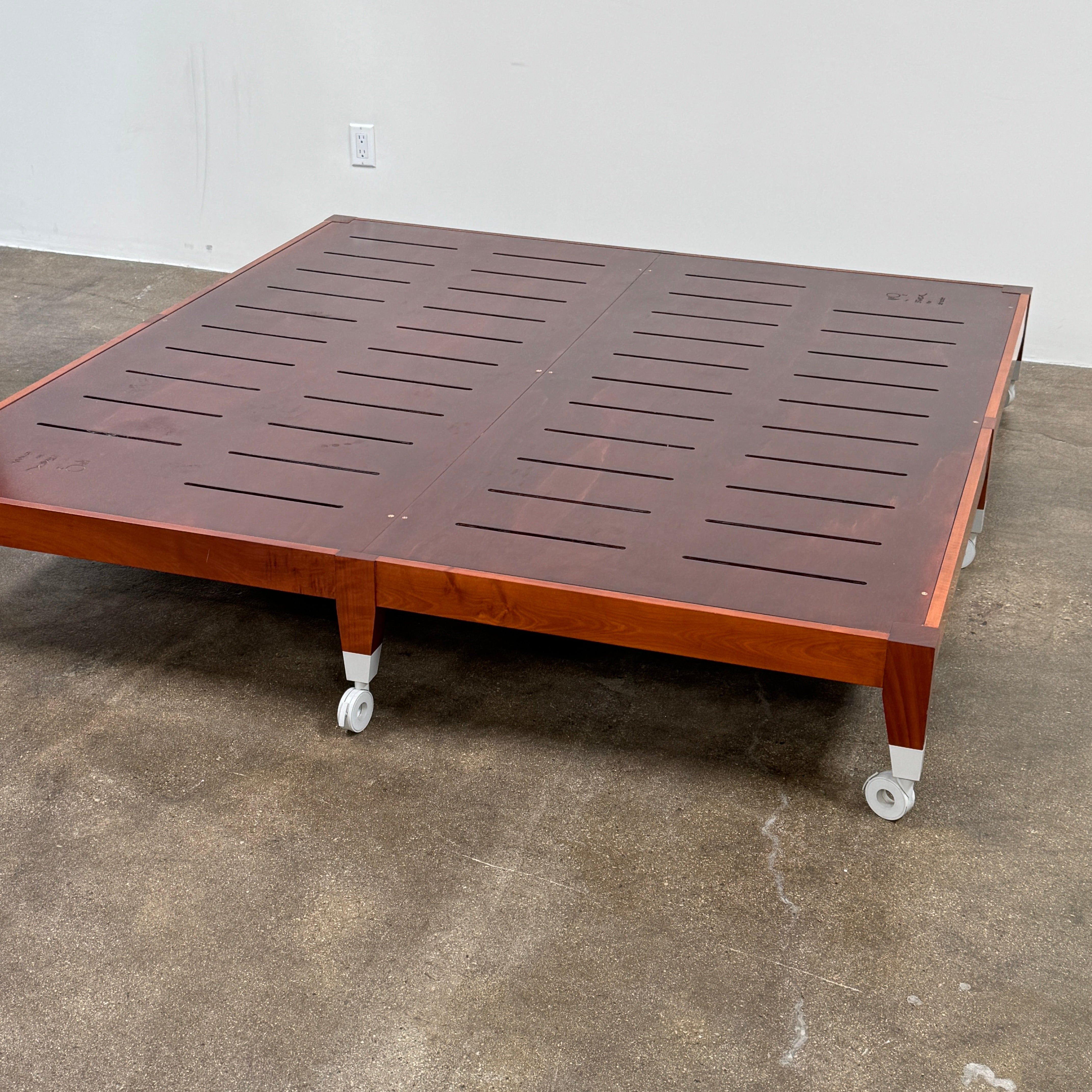 The Driade Neoz European King Size Bed by Driade, featuring wooden slats, wheels, and a white fabric headboard, stands on a concrete floor against a plain white wall.