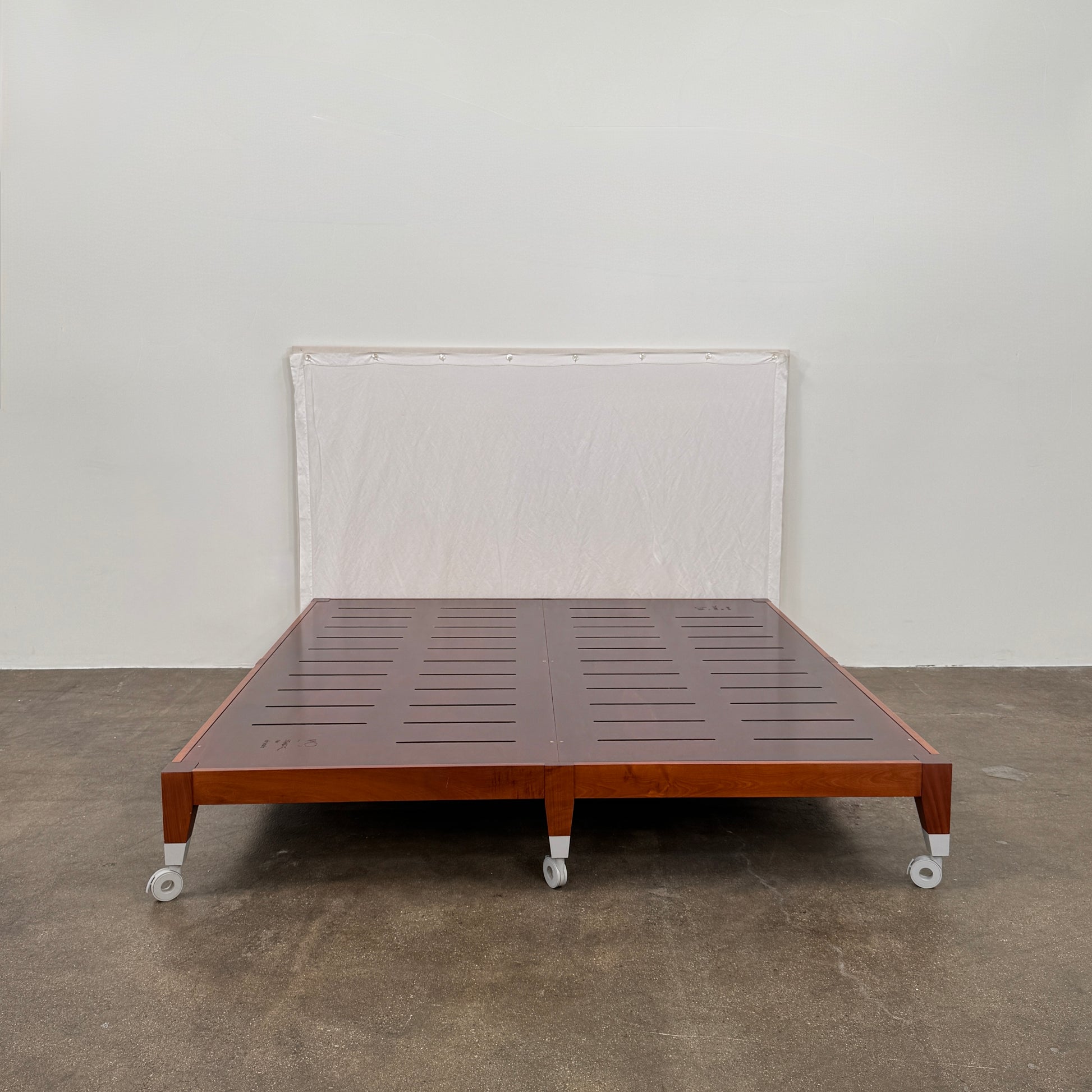 The Driade Neoz European King Size Bed by Driade, featuring wooden slats, wheels, and a white fabric headboard, stands on a concrete floor against a plain white wall.