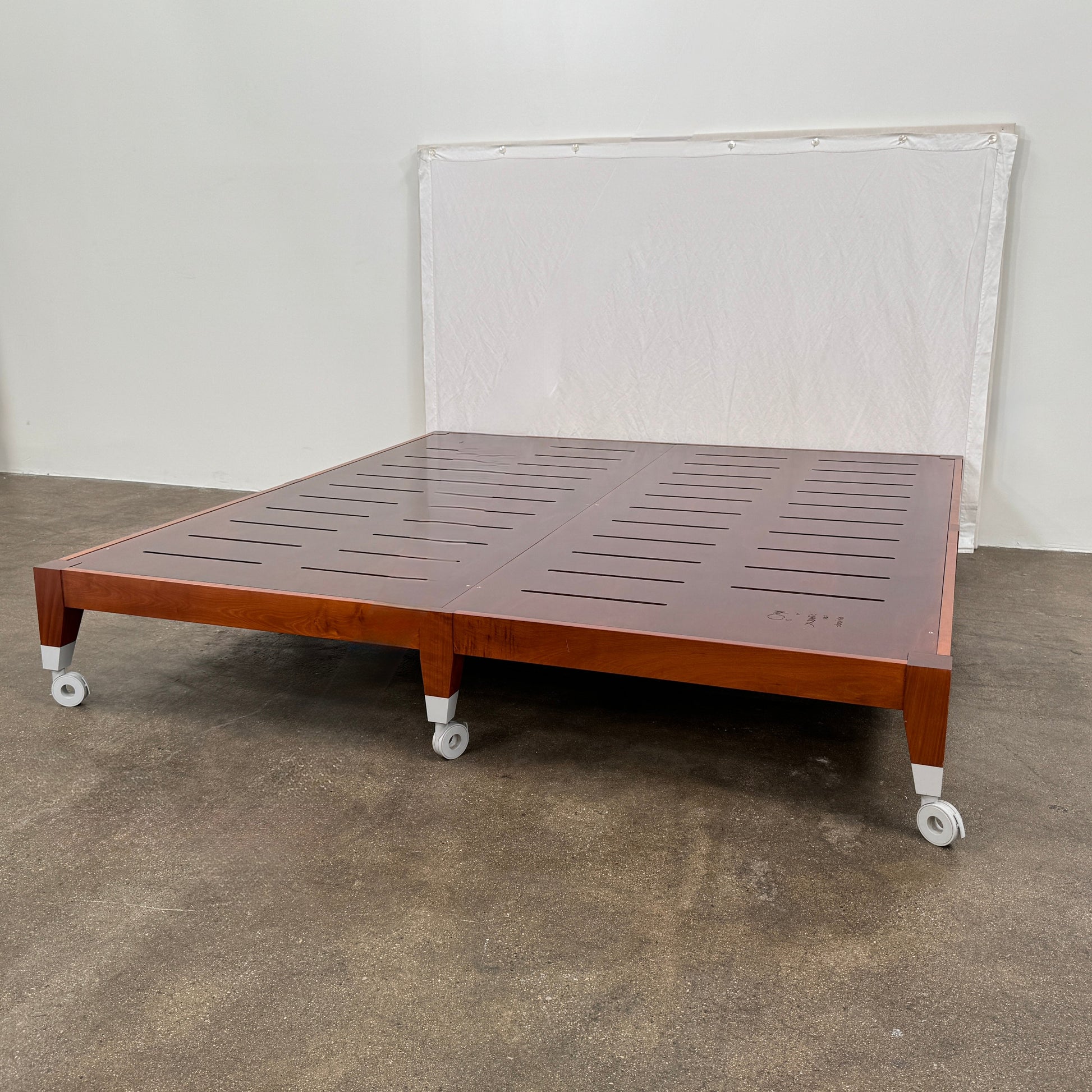 The Driade Neoz European King Size Bed by Driade, featuring wooden slats, wheels, and a white fabric headboard, stands on a concrete floor against a plain white wall.